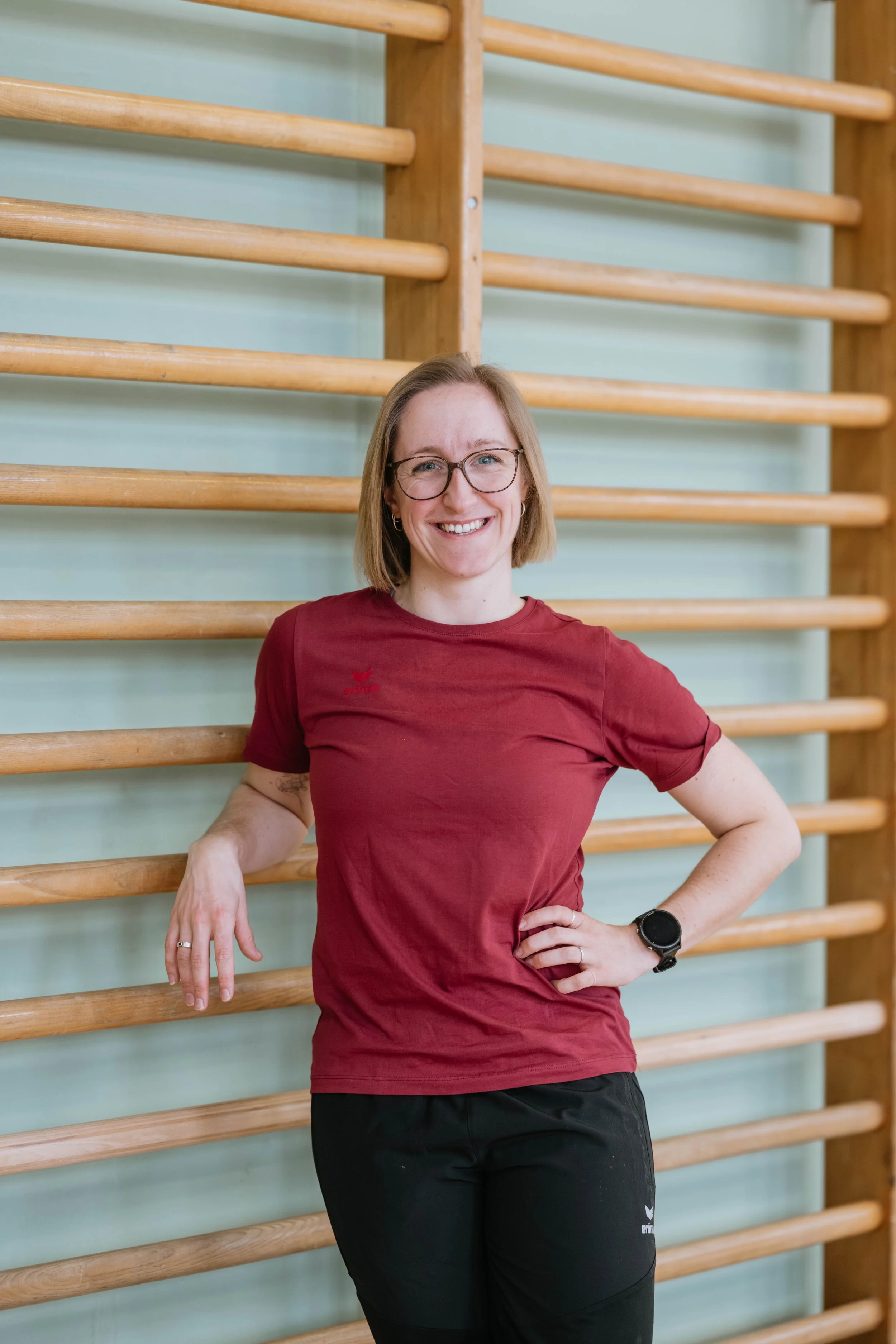 Frau mit Brille und einem Lächeln, trägt eine rote Sport T-Shirt und schwarze Sporthose, steht vor einer Holzturnstange in einer Turnhalle.