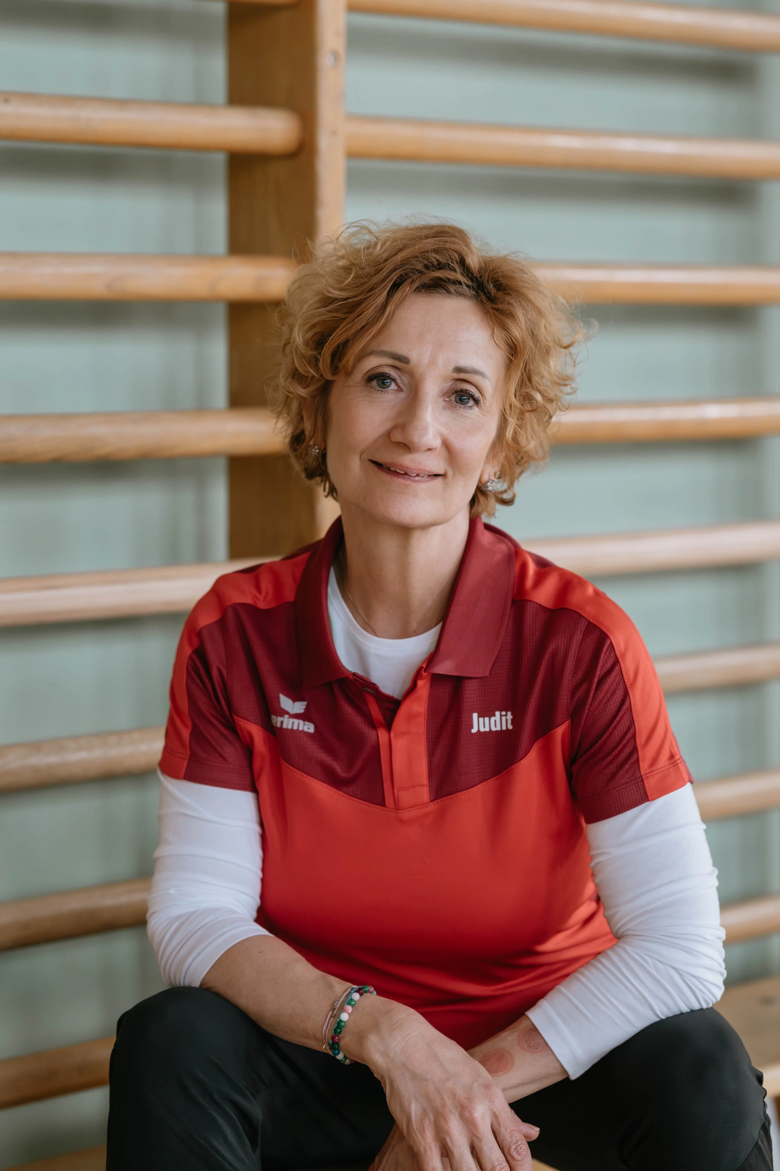 Eine ältere Frau mit kurz lockigem, rotem Haar sitzt auf einer Bank in einer Turnhalle. Sie trägt eine rote Sport-Uniform mit dem Namen 'Judit'.