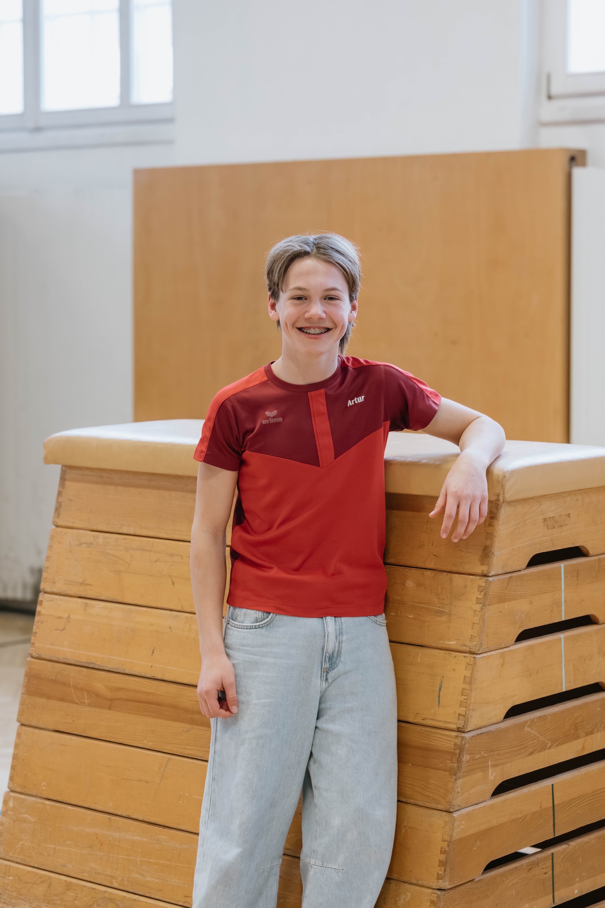 Ein junger Junge mit braunem Haar, lächelnd, trägt ein rotes Sportshirt mit Namen "Artur" und hellblaue Jeans, lehnt an turnen Matten in einer Turnhalle.