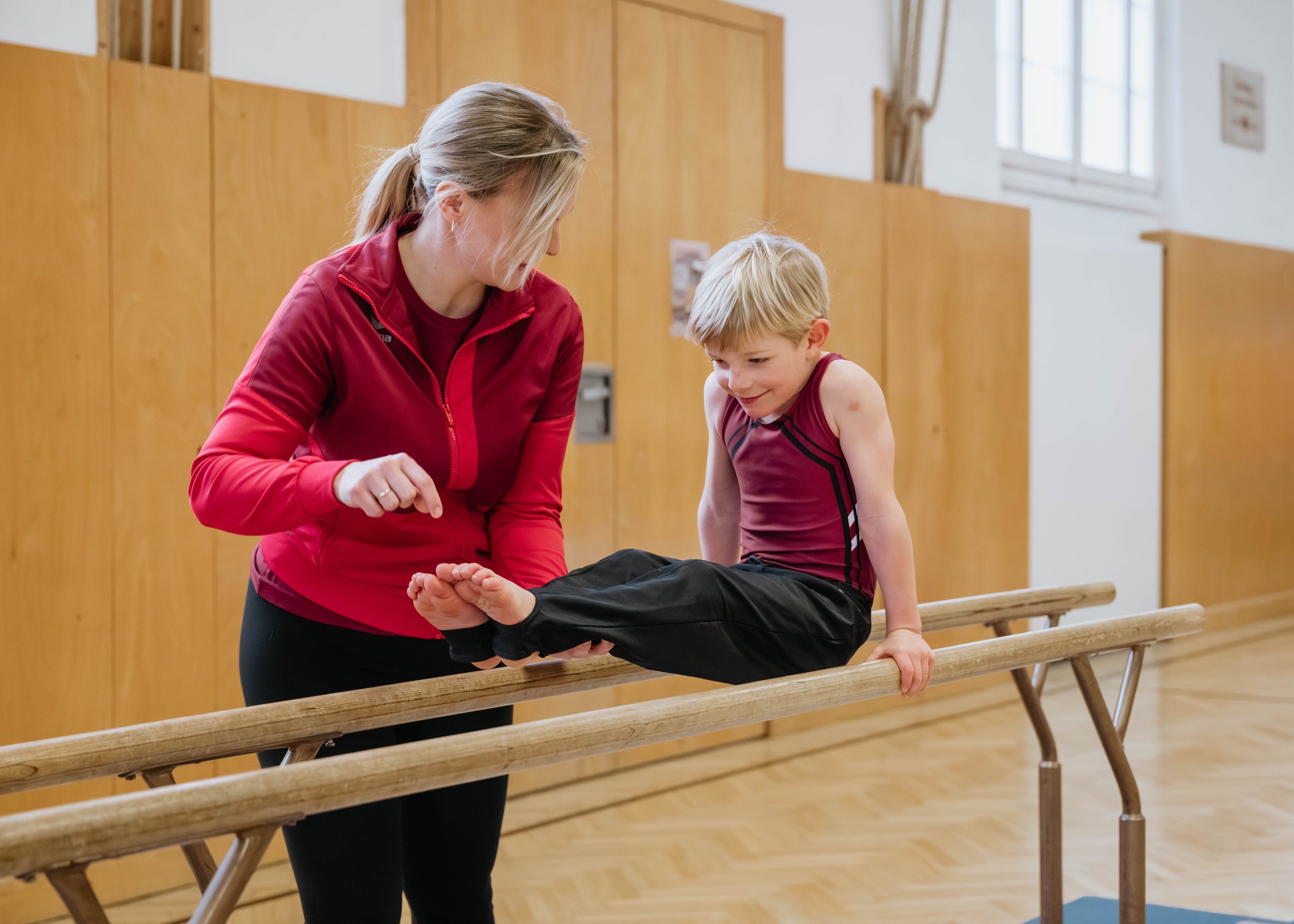 Eine Trainerin hilft einem Jungen beim Gleichgewichtstraining auf einer Barren im Turnraum.