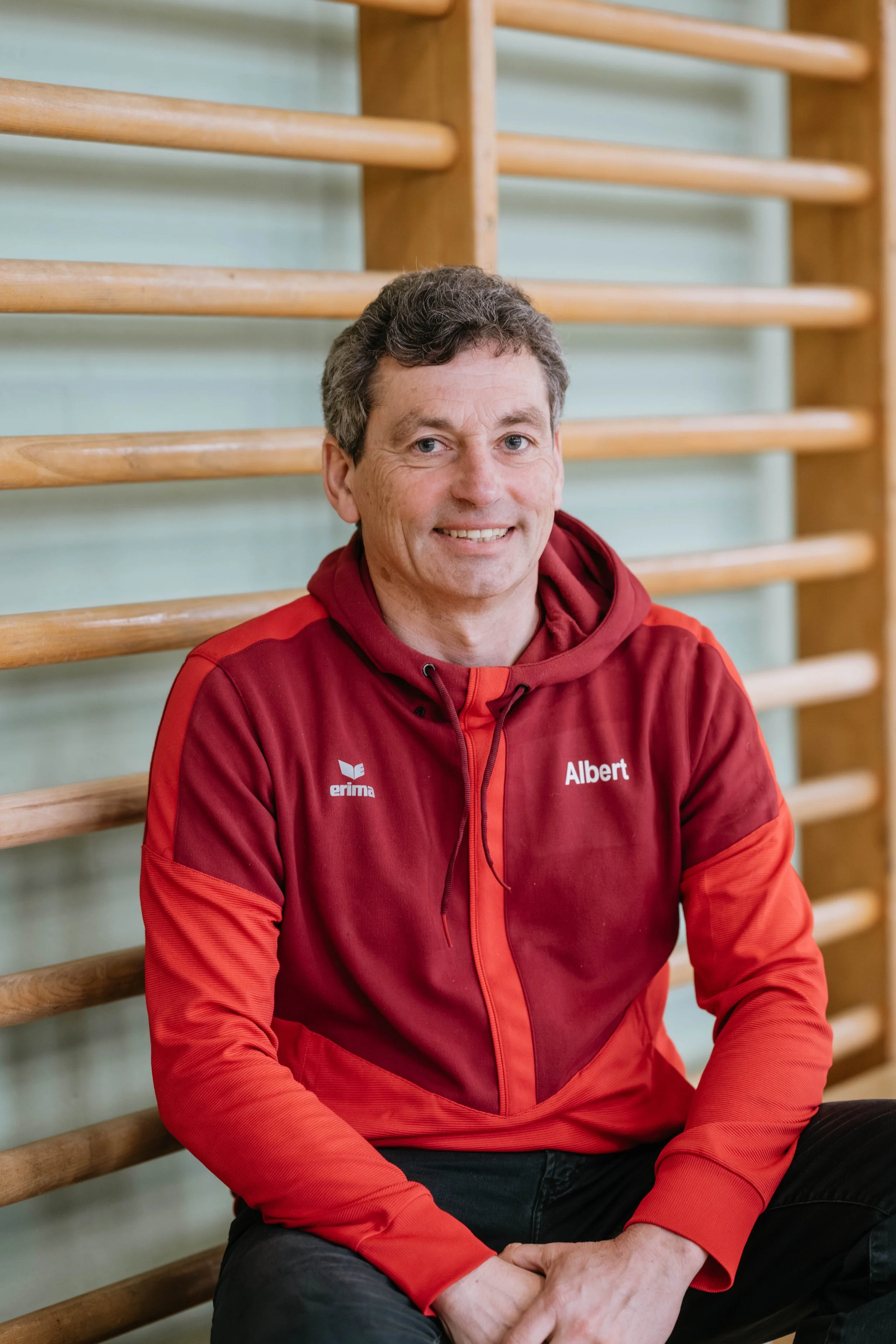 Mann in rotem Sportsjacke sitzend vor einer Sprossenwand in einer Turnhalle.