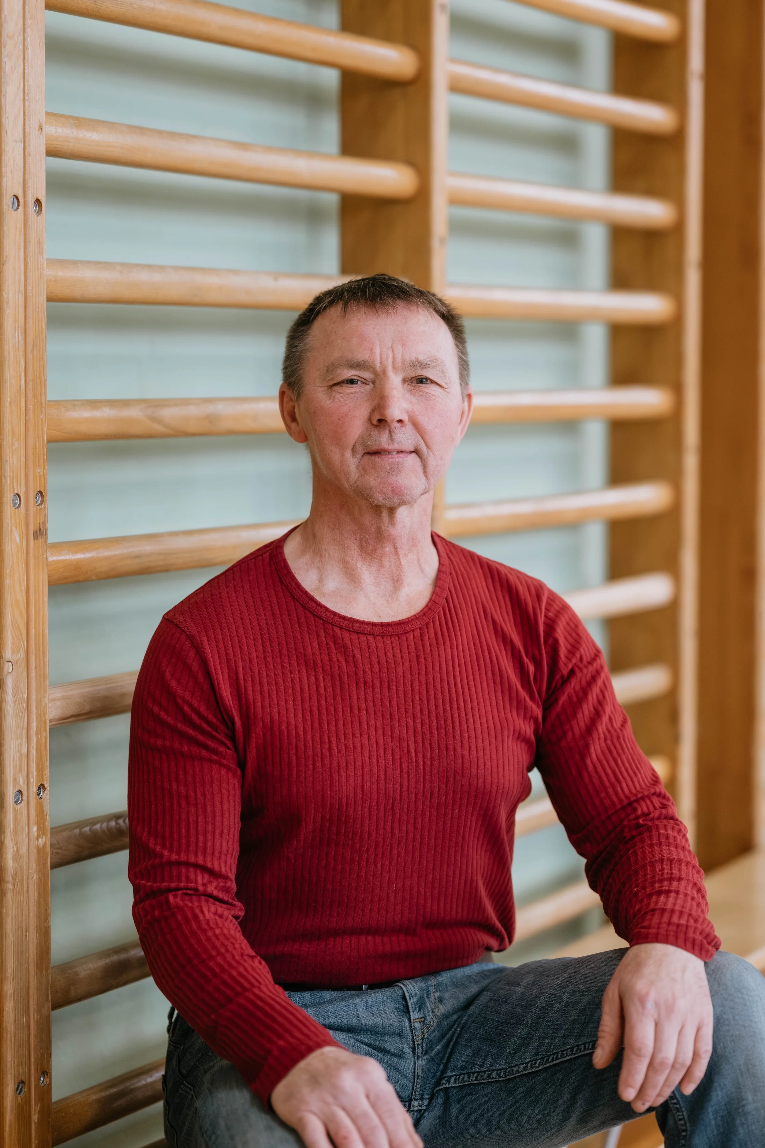 Ein älterer Mann sitzt an einer Wand mit Turngeräten und blickt in die Kamera. Er trägt ein rotes, langärmliges Hemd und Jeans.