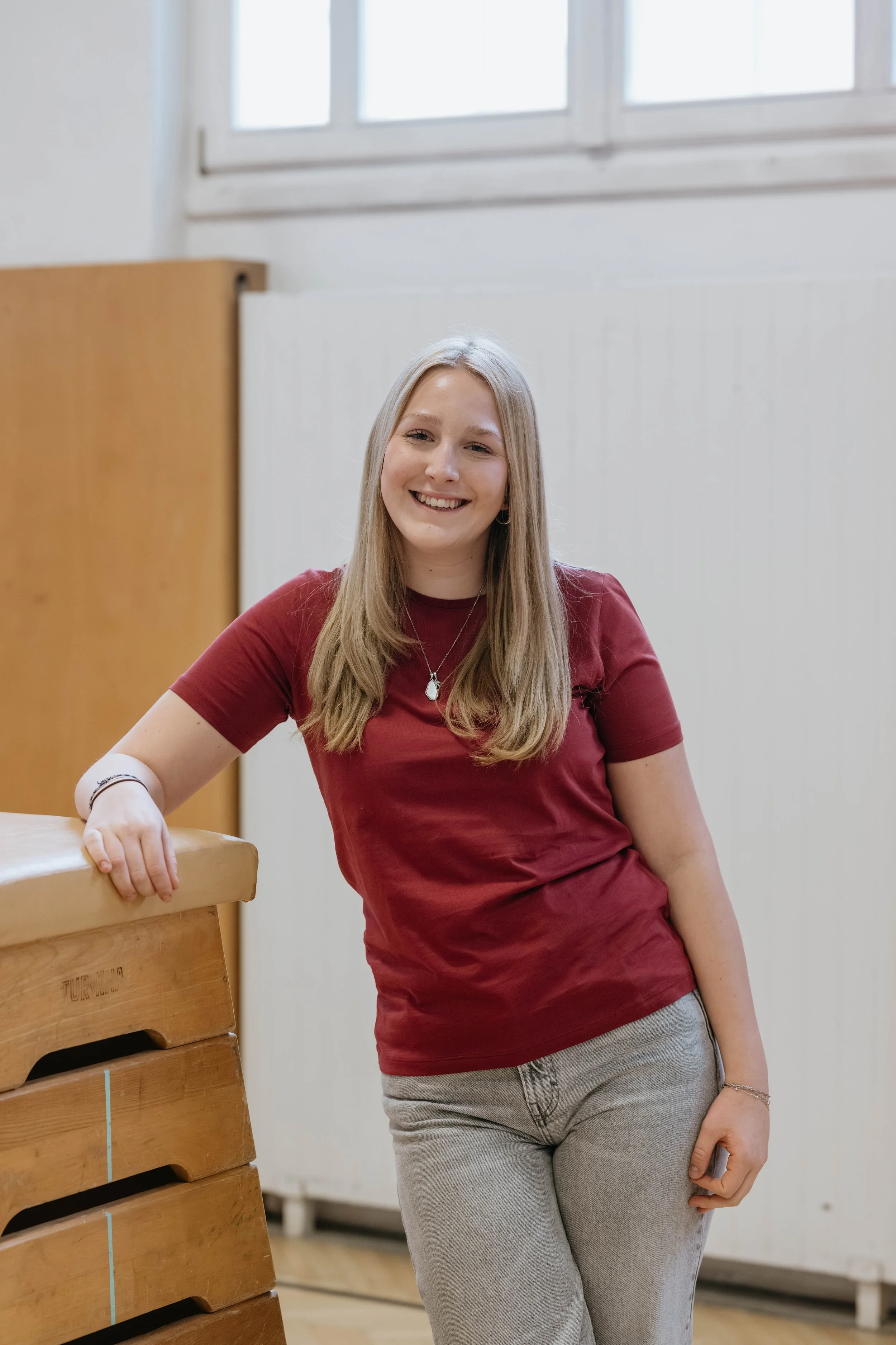 Lächelnde junge Frau in rotem T-Shirt und grauen Hosen steht in einem Raum mit weißen Wänden, neben einem Holzpult.