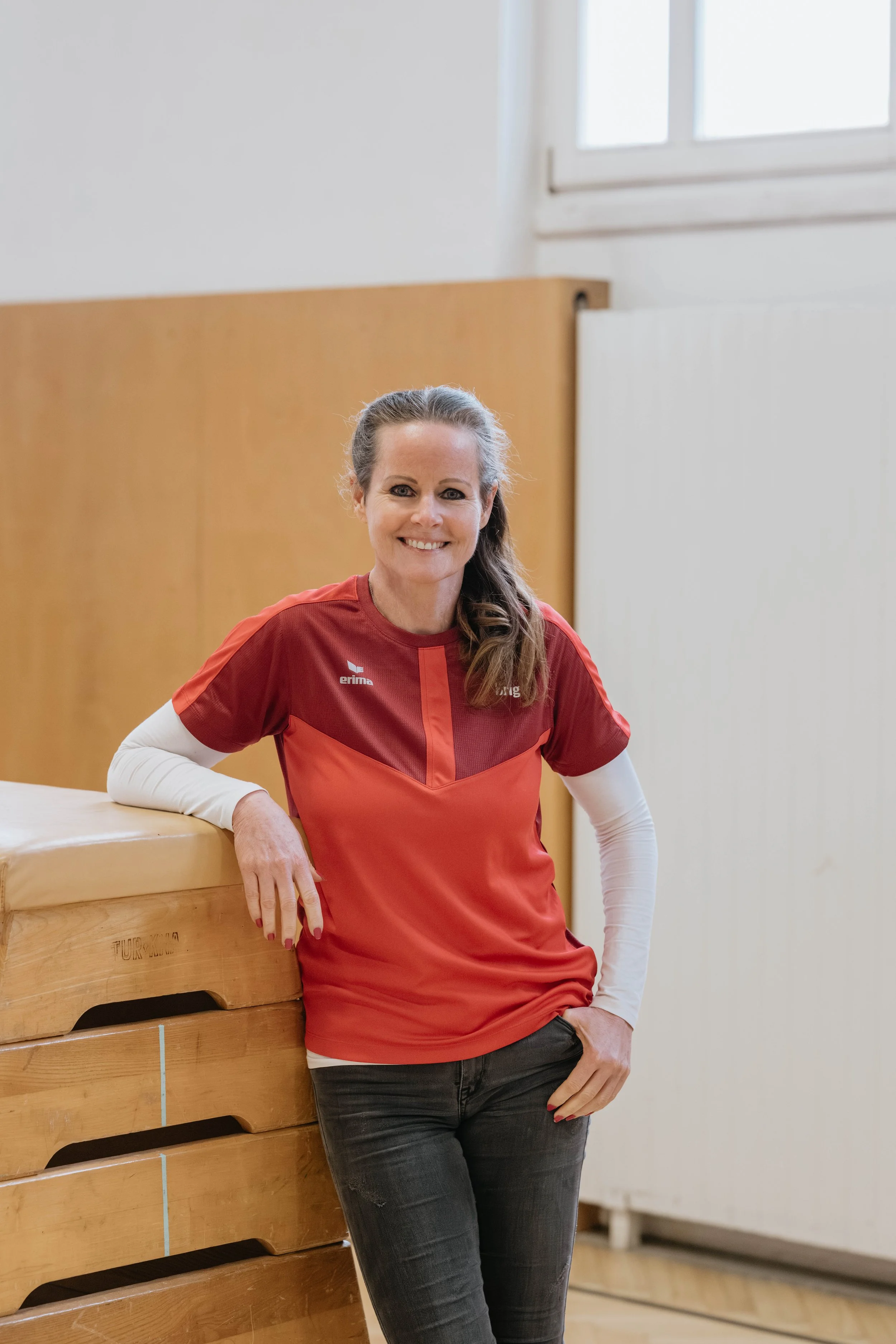 Eine lächelnde Frau in rotem Sportshirt lehnt an einem Holzkasten in einer Turnhalle.