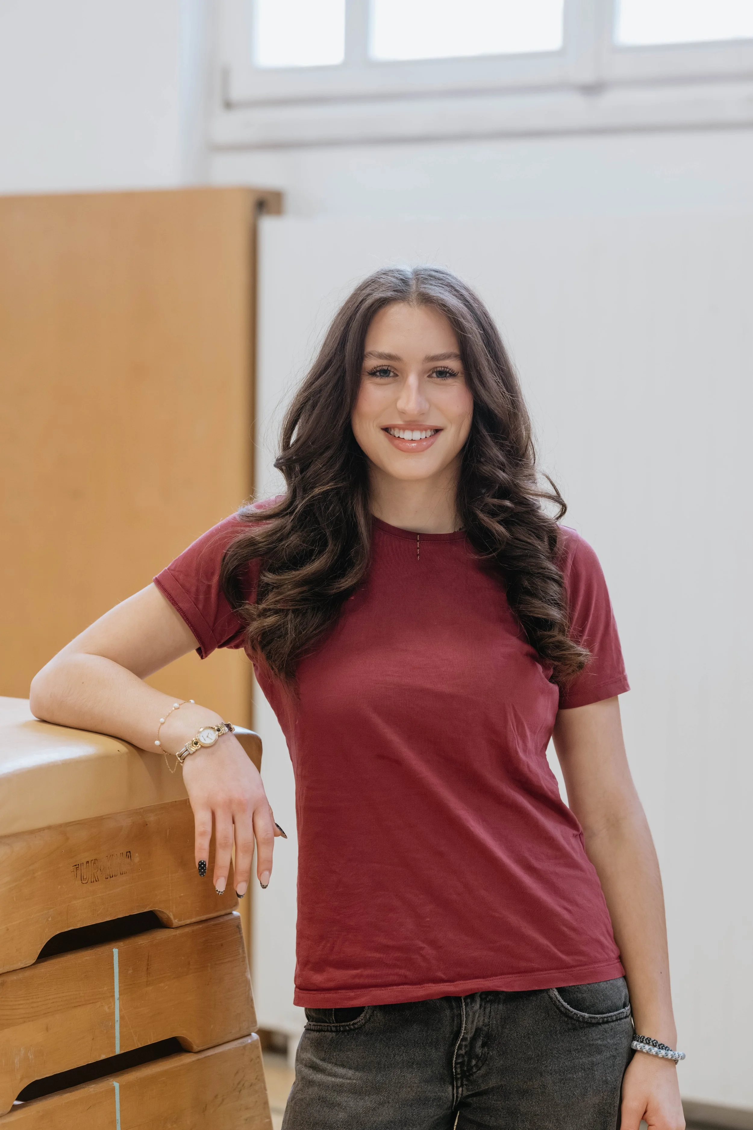 Ein junge Frau mit langem braunem Haar, lächelnd, trägt ein rotes T-Shirt, steht neben einem Holzgerät im Innenraum.