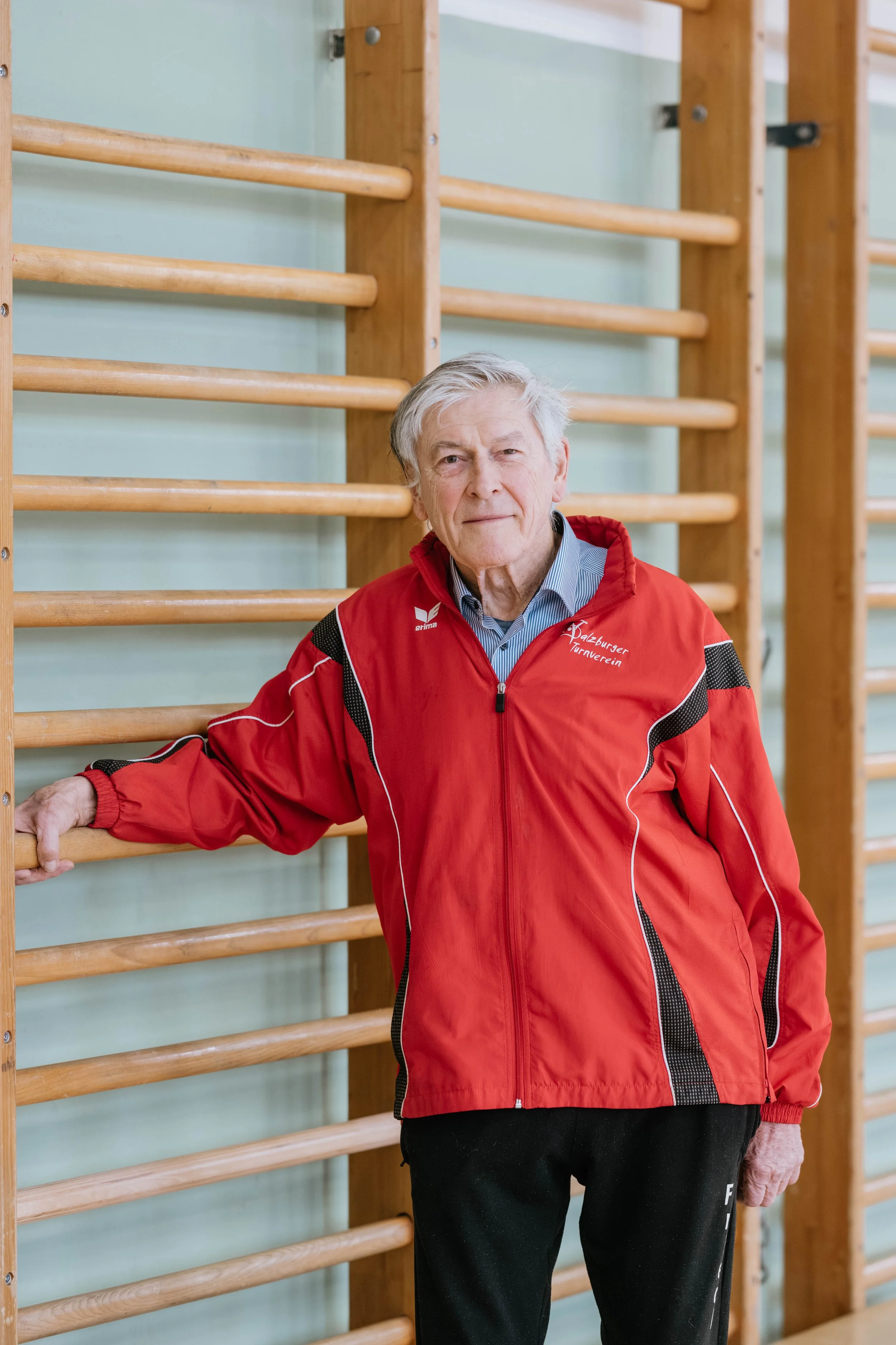Älterer Mann in roter Turnjacke steht in Turnhalle vor Holzreckwand, blickt in die Kamera.