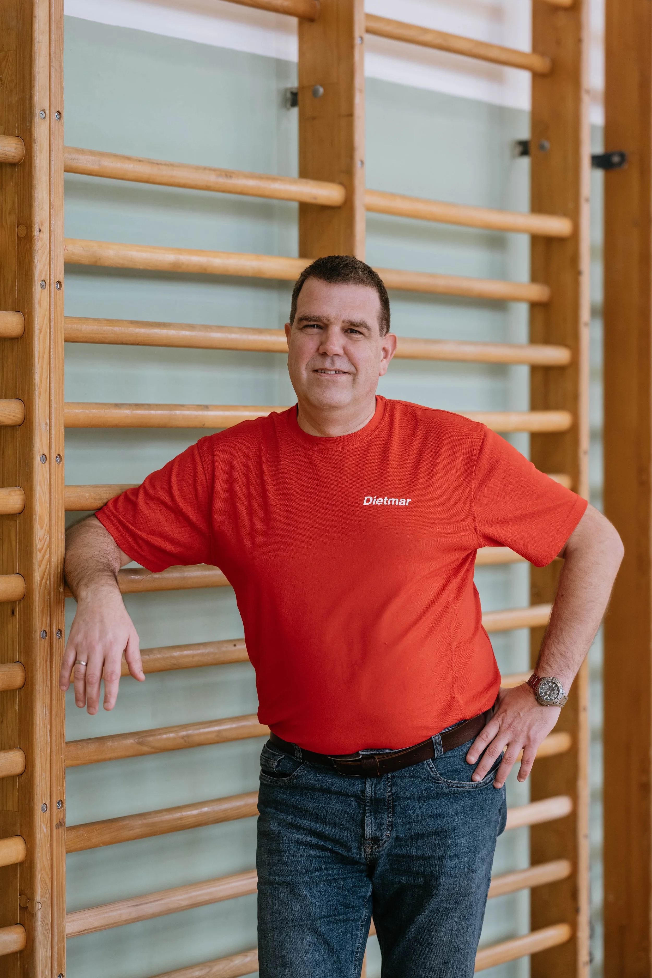 Ein Mann trägt ein rotes T-Shirt mit dem Schriftzug ‚Dietmar‘ und steht in einem Fitnessraum vor einer Holzturnstange. Er lehnt mit einem Arm an der Turnstange und lächelt, während er die andere Hand auf die Hüfte legt.