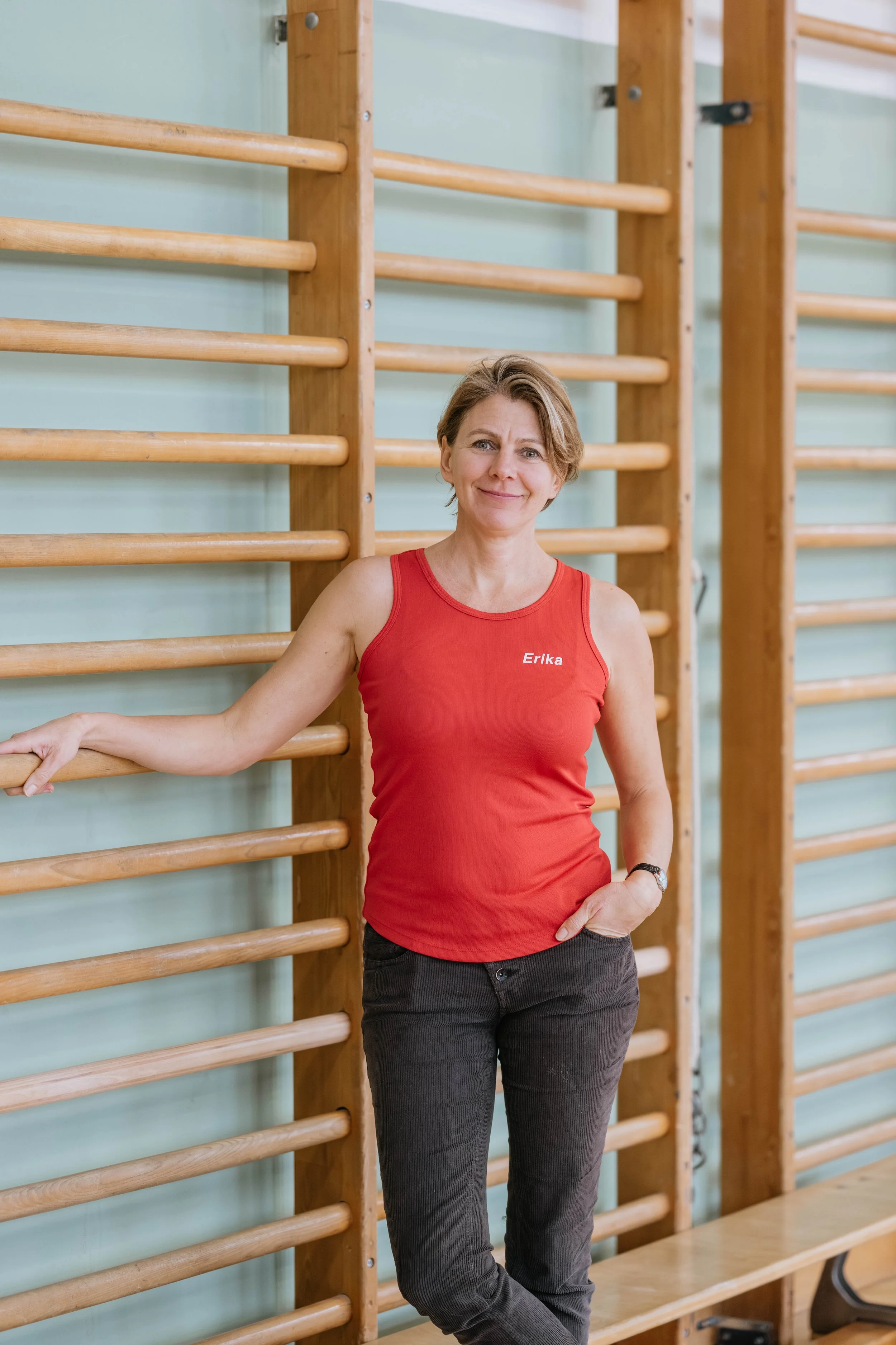 Eine Frau mit kurzen braunen Haaren und einem roten Top mit der Aufschrift ‚Erika‘ steht vor einer Holzwand im Fitnessraum, lehnt an einem Horizontalbalken.