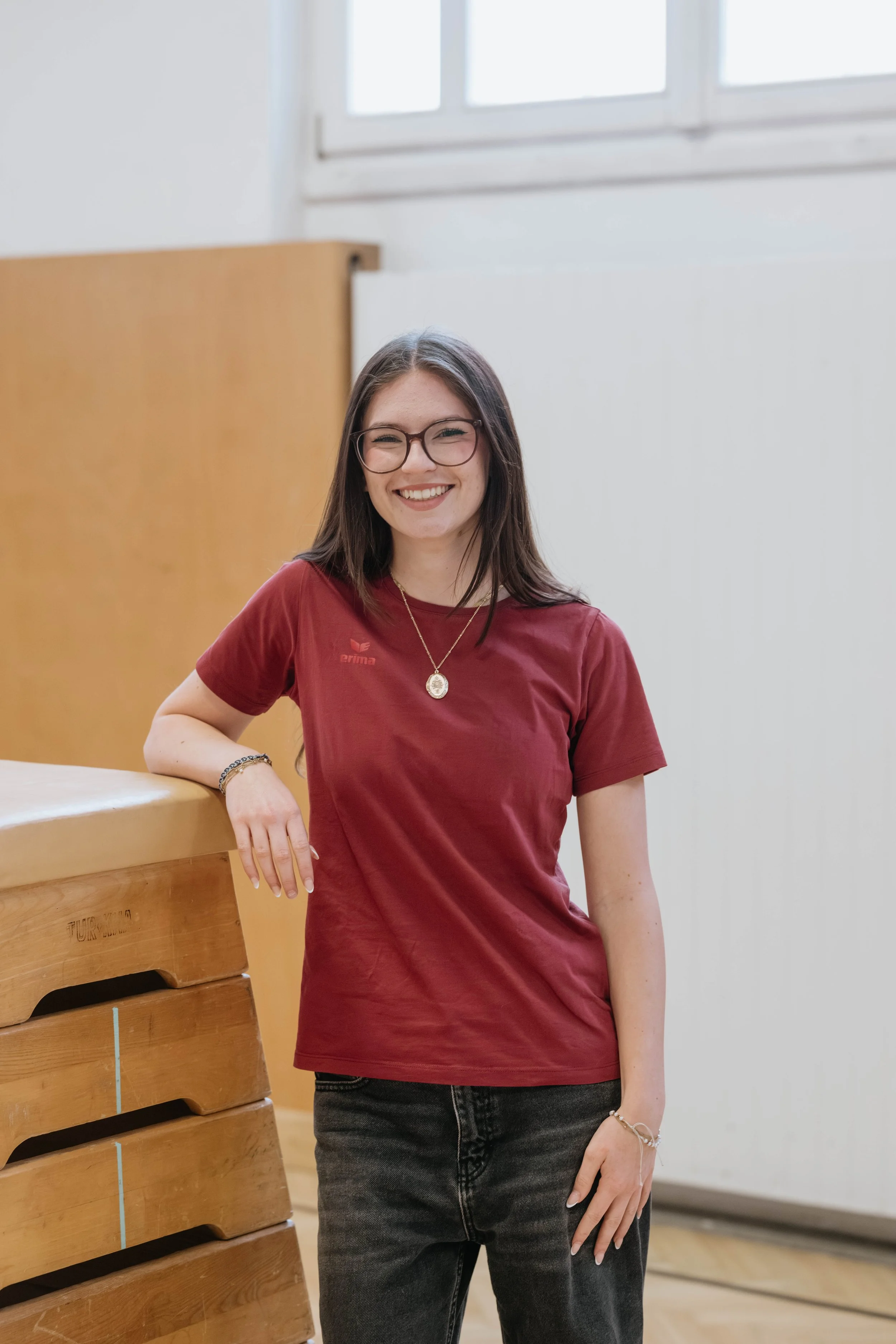 Lächelnde junge Frau mit Brille, dunklem Haar, rotem T-Shirt und dunklen Jeans, die an einer Turnmatte im Fitnessstudio steht.