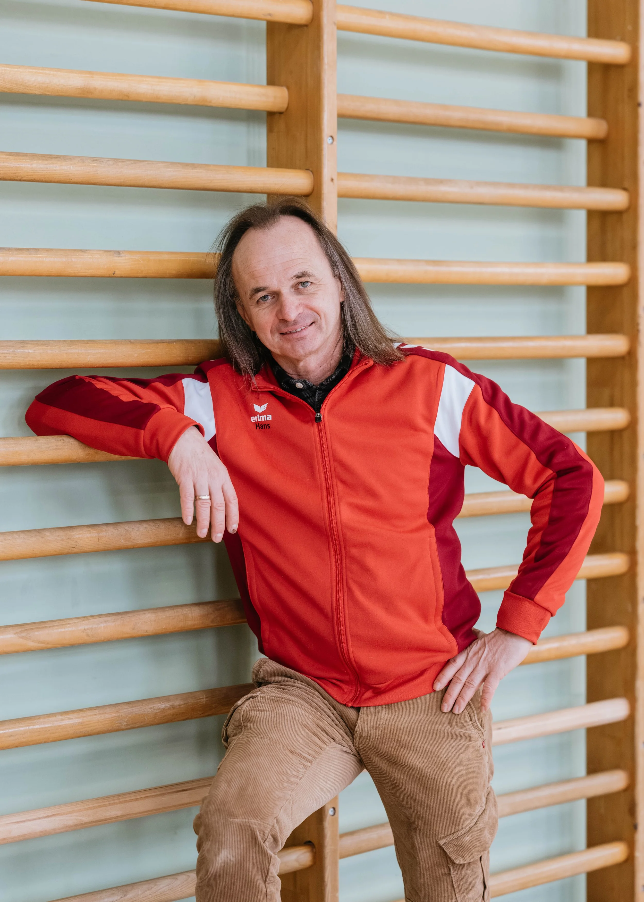 Ein Mann in rotem Sportjacke lehnt an einer Holz-Trainingswand in einer Turnhalle.