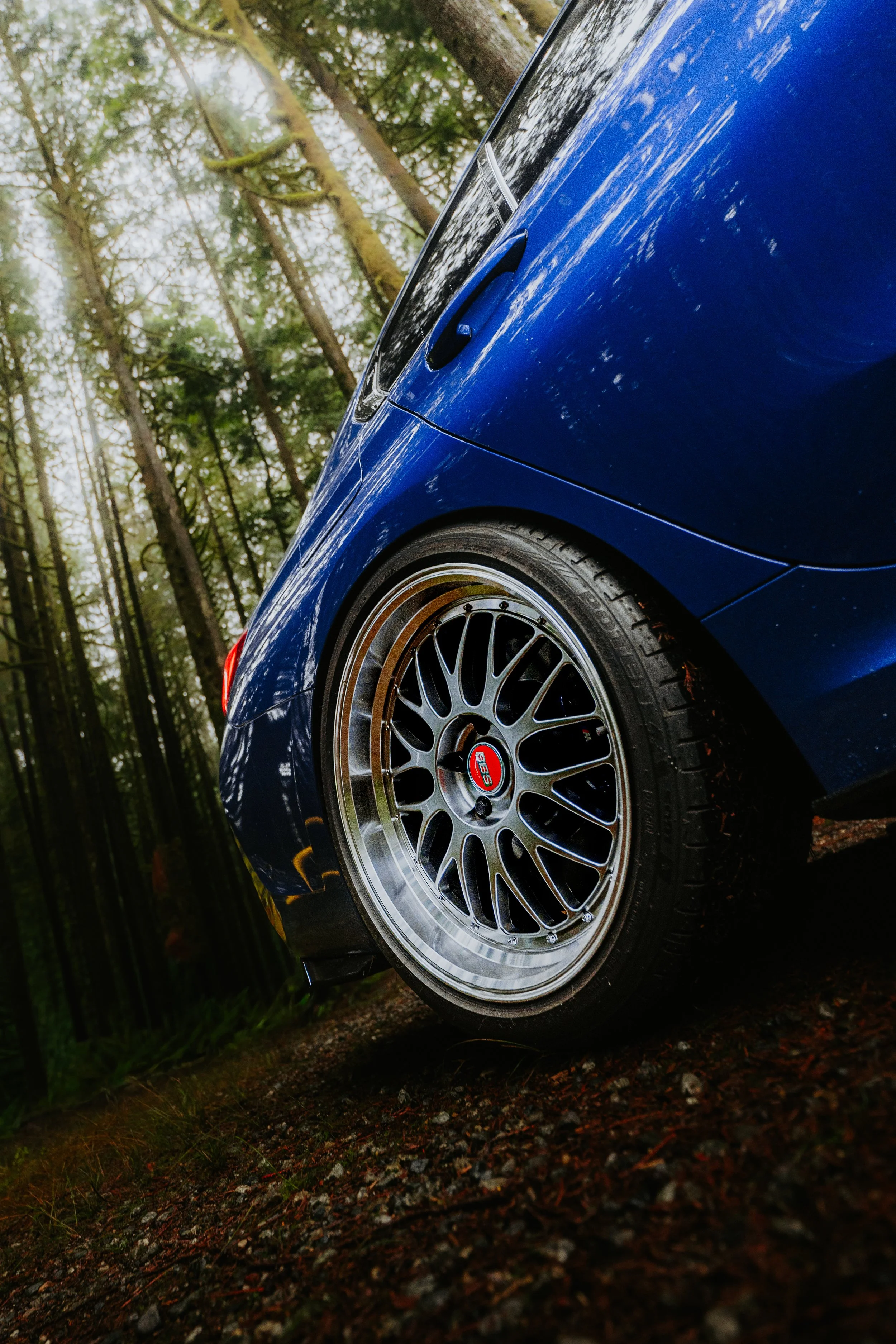 Close-up of a blue car with aftermarket wheels, parked in a wooded area with tall trees.