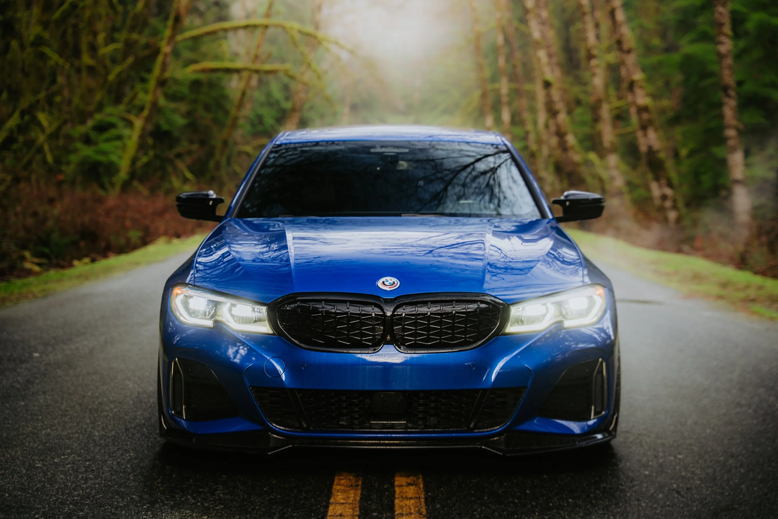 A blue car on a narrow road through a forested area with trees and greenery.