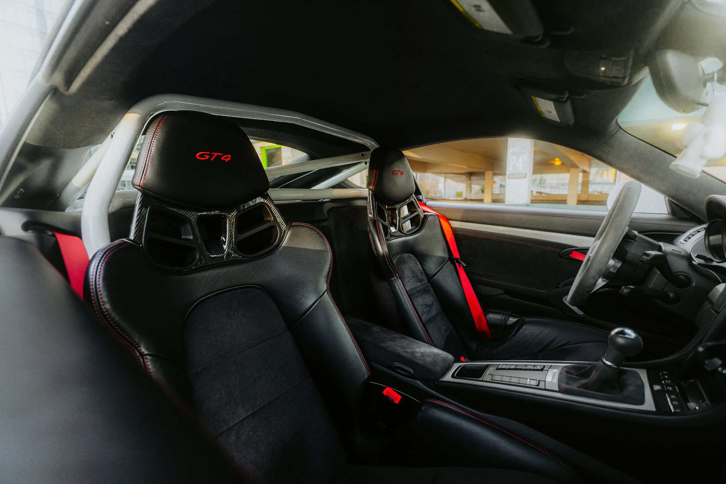 Inside view of a Porsche 911 GT4 race car with two racing seats, a roll cage, and a gear shifter in the center console, parked in a parking garage.