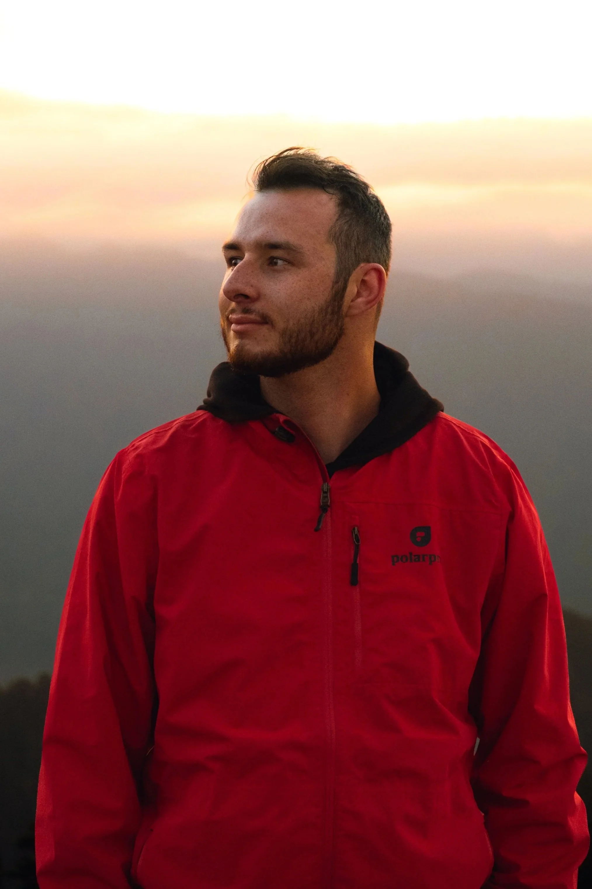 A young man with short dark hair and a beard wearing a red jacket with black accents, standing outdoors during sunset or sunrise.