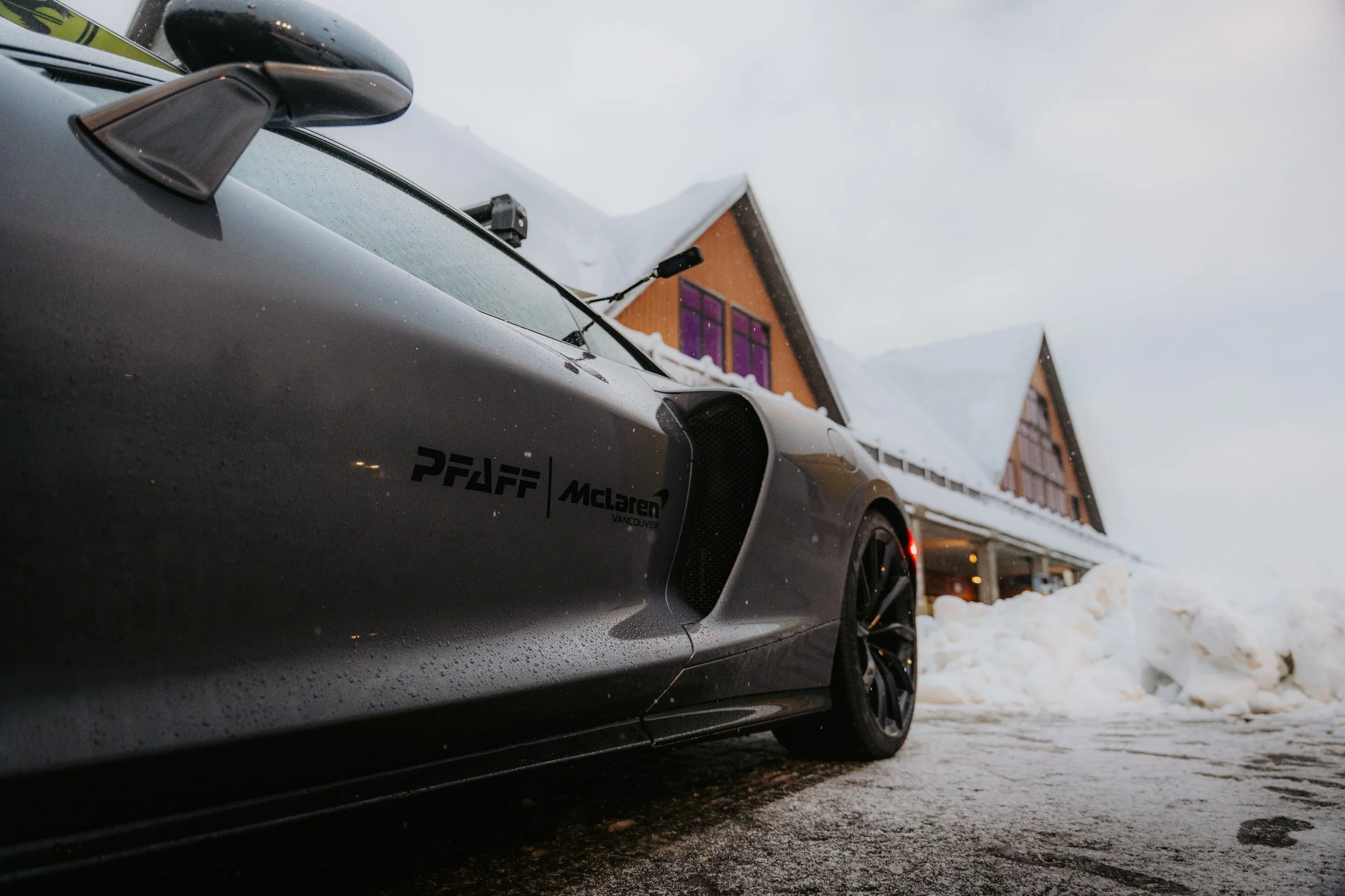 A gray McLaren sports car with water droplets on its surface, parked outside a building with snow on the ground and snow-covered rooftops in the background.