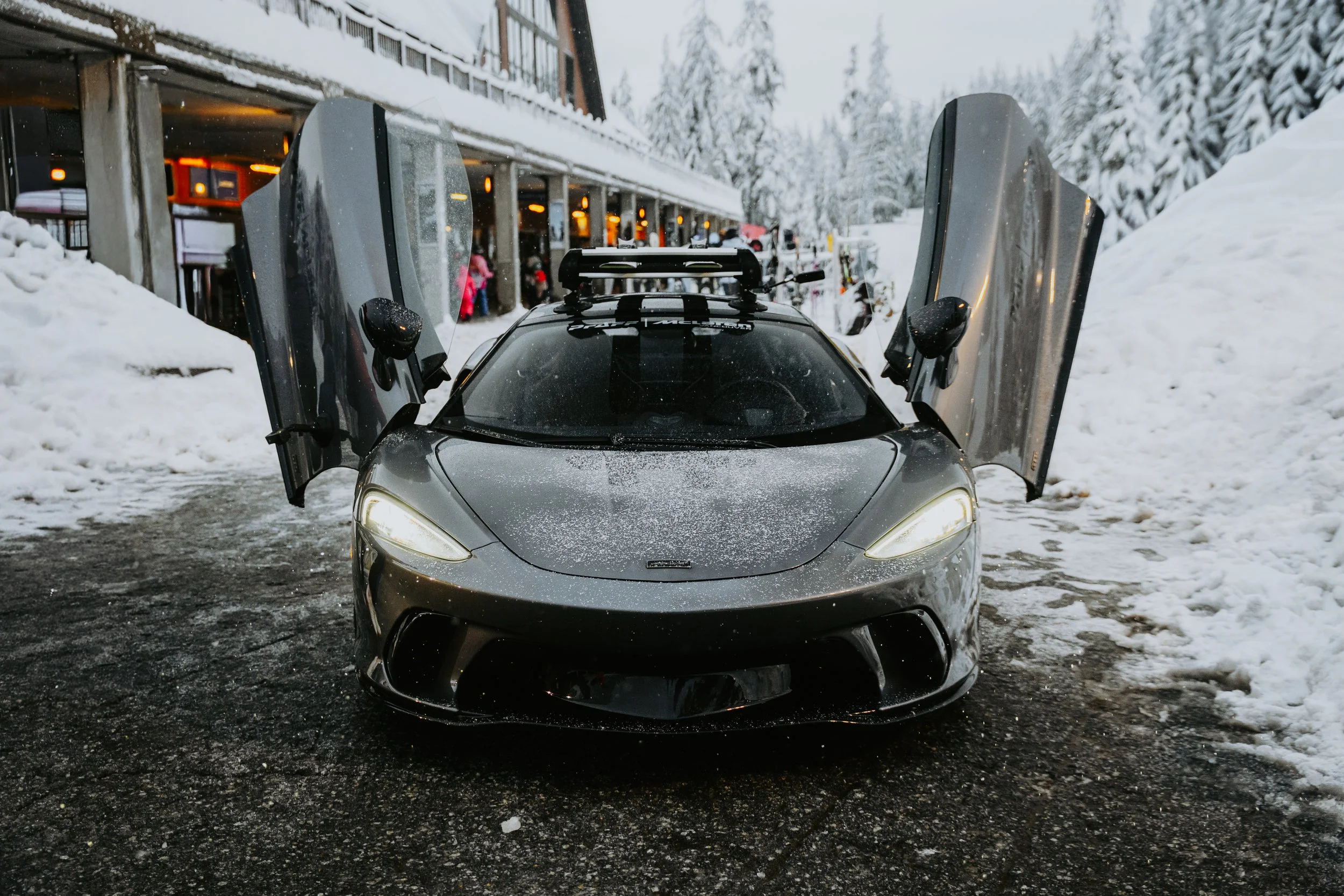 A sleek, silver sports car with its butterfly doors open, parked on a snowy parking lot in front of a building with large windows and warm lighting. Snow-covered trees surround the scene.