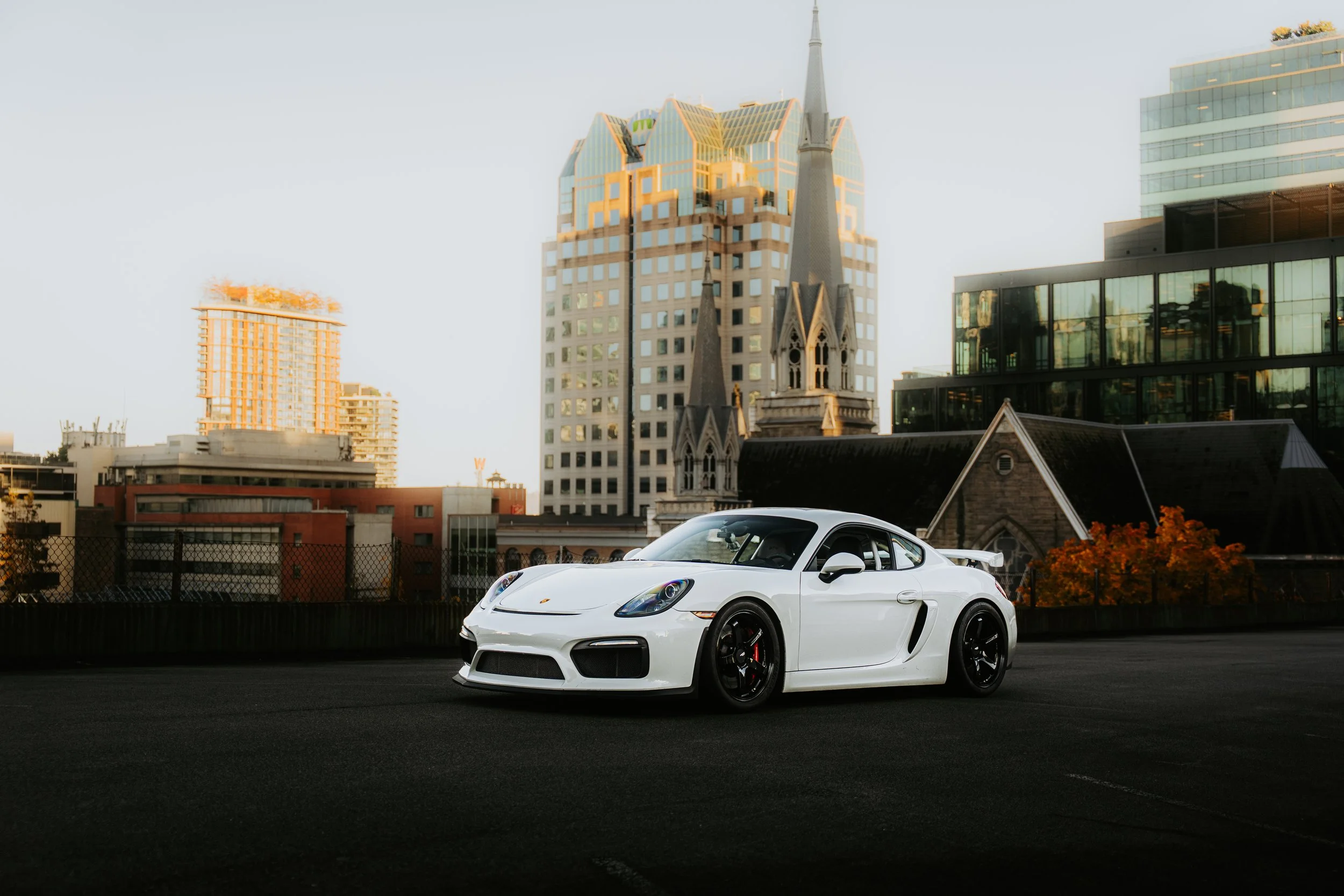 White sports car parked on a city rooftop with tall buildings and a church in the background during sunset.