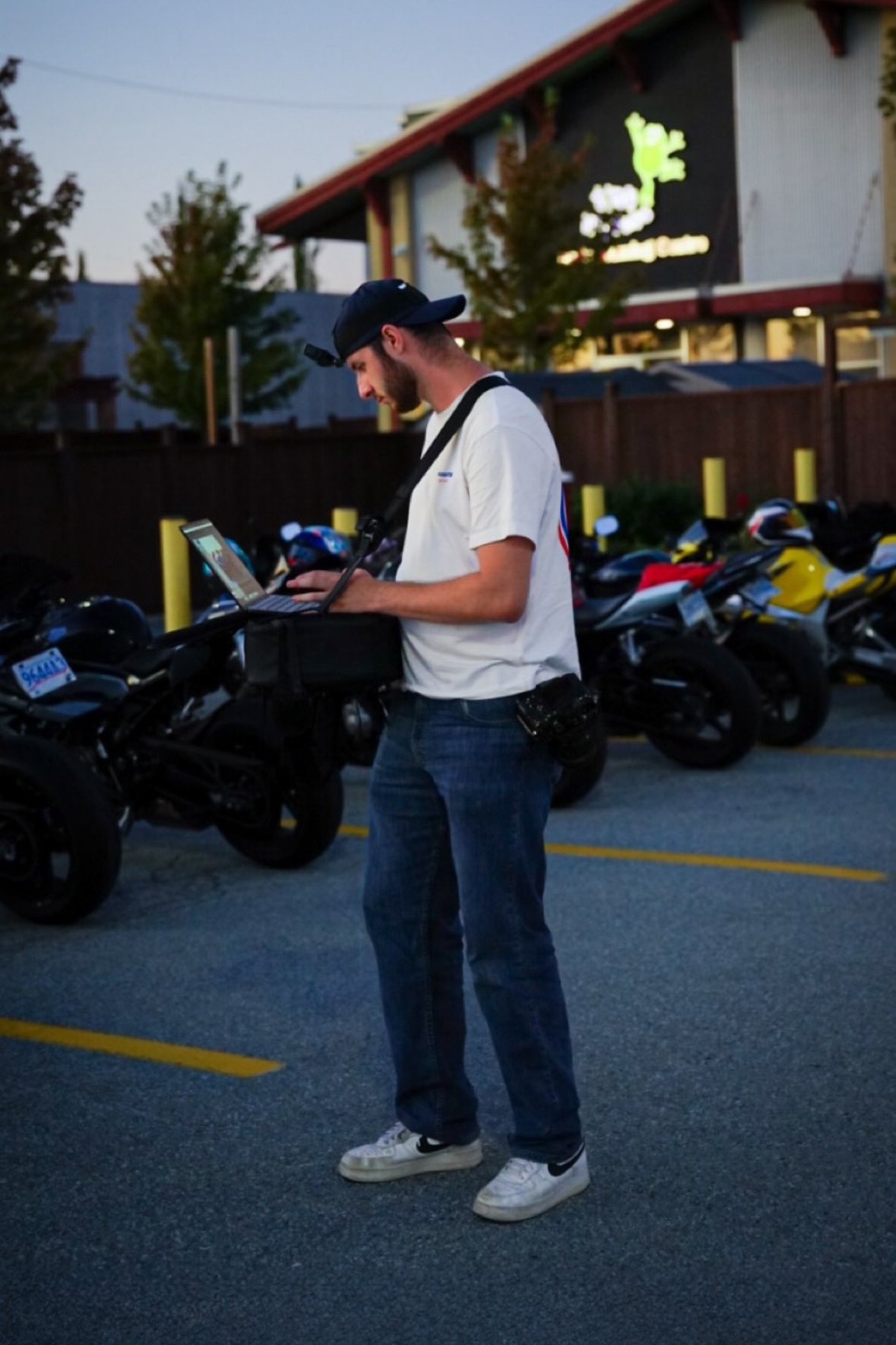 A man standing in a parking lot using a laptop on a portable desk. He is wearing a white t-shirt, blue jeans, white sneakers, and a backwards cap. Several motorcycles are parked nearby, and a building with a brightly lit sign is visible in the background during dusk.