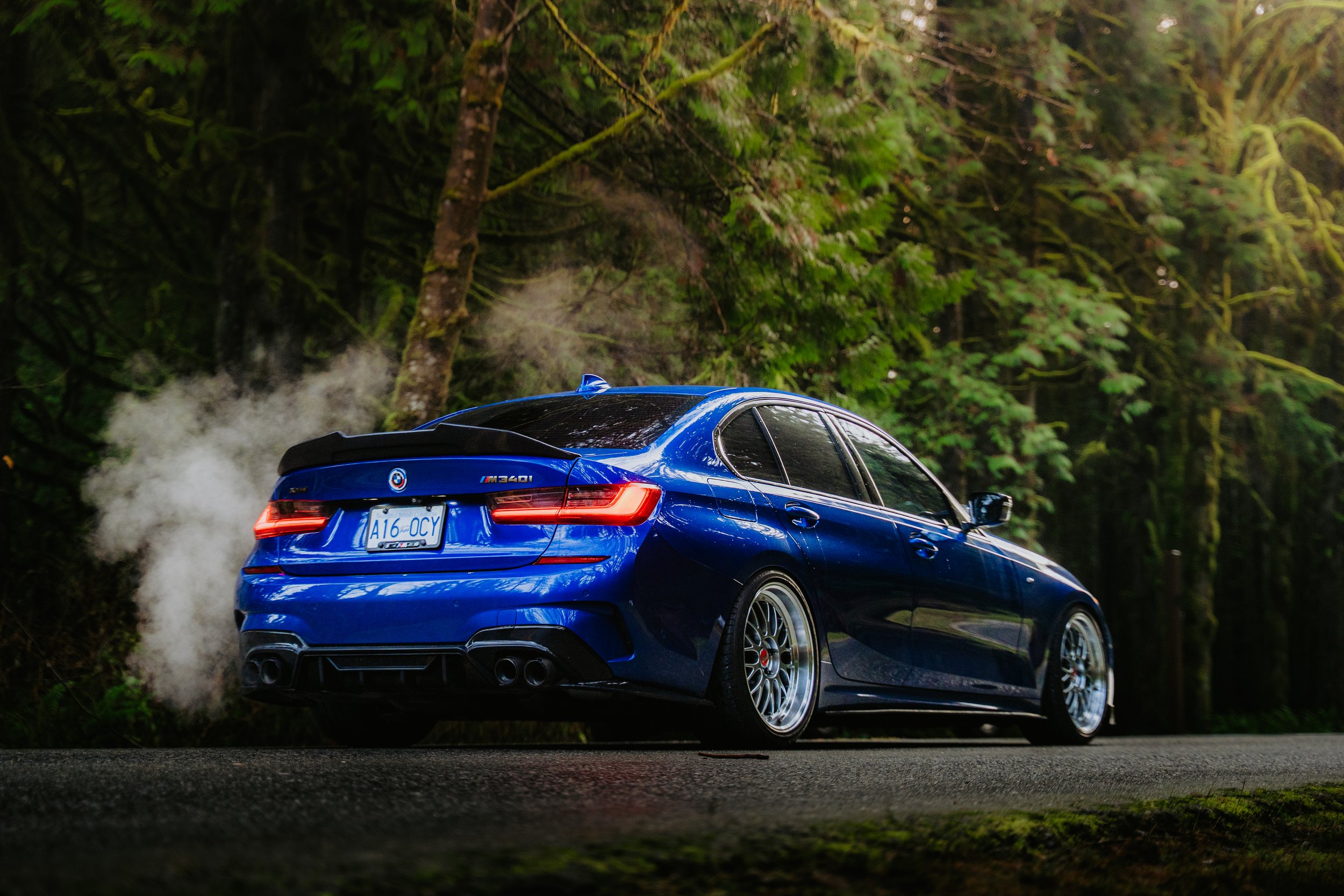 A blue BMW M340i driving on a forest road with trees and greenery in the background, emitting smoke from the exhaust.