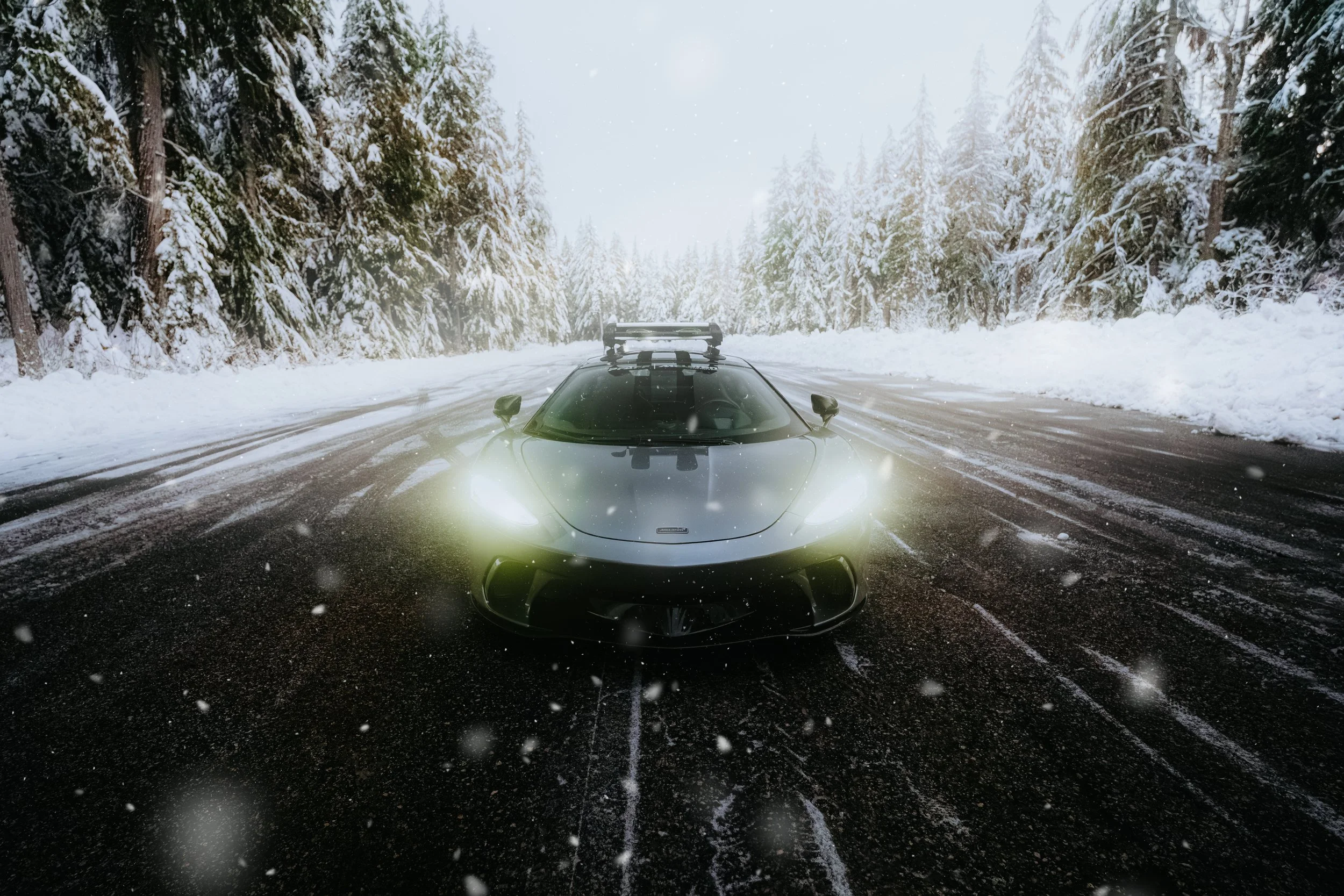 A sleek, black sports car with its headlights on, driving on a snow-covered road surrounded by snow-laden evergreen trees in a winter landscape.
