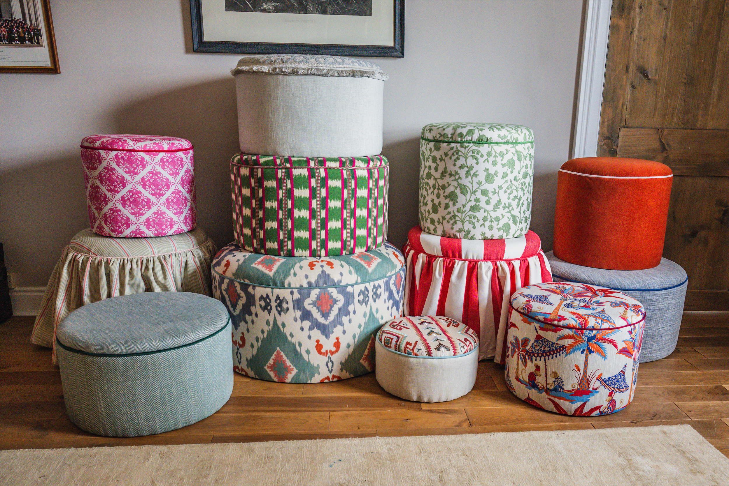A collection of colorful, patterned upholstered ottomans and stools arranged in a living room corner against a wall with framed pictures. The ottomans feature various fabrics including florals, stripes, and abstract designs.