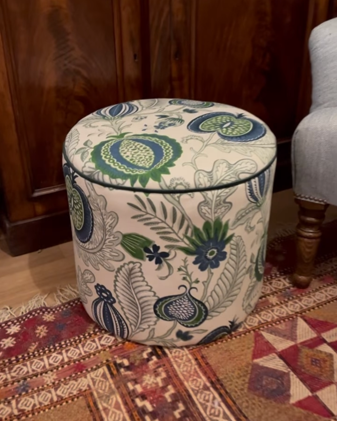 A round, upholstered ottoman with a floral and fruit pattern in blue, green, and beige colors, placed on a patterned rug with a wooden panel wall and part of a gray armchair visible in the background.