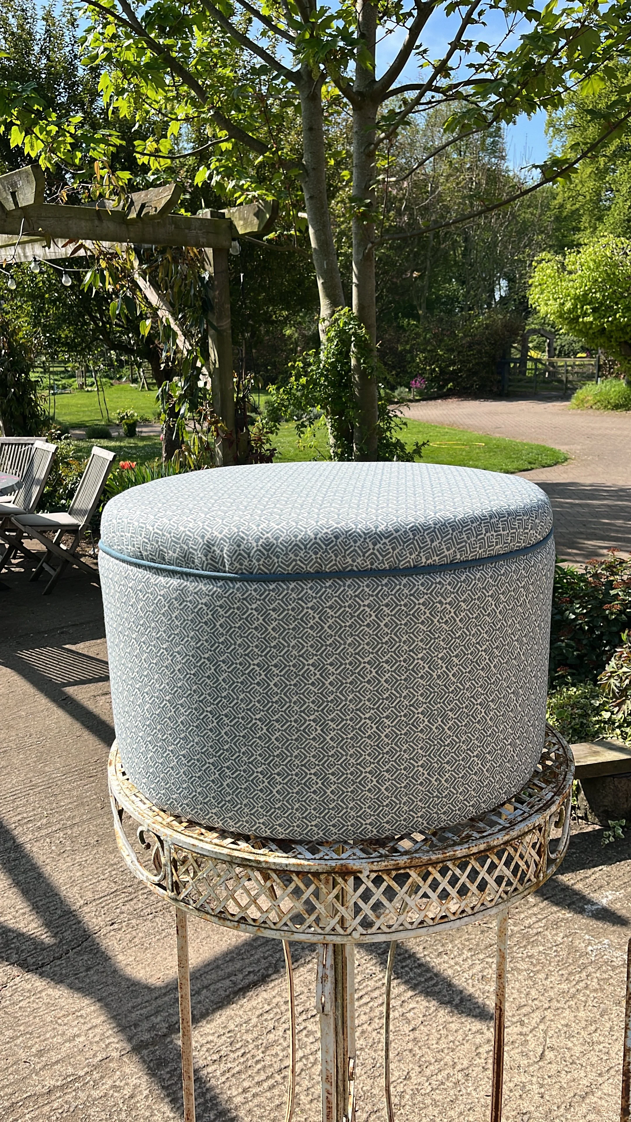 A round outdoor table with a large, round, decorative fabric-covered cushion on top, placed on a metal stand in a garden setting with trees, shrubs, and a gravel path.