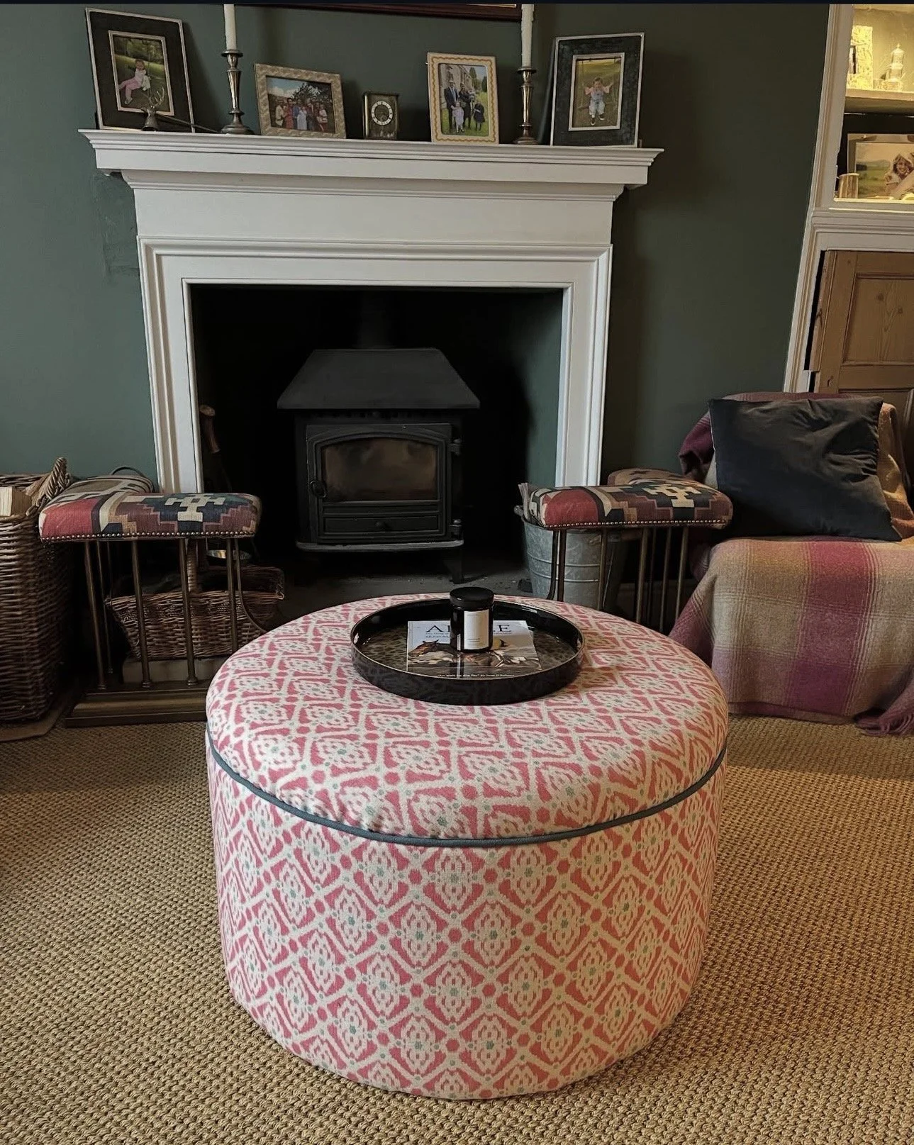 Living room with a white fireplace mantle decorated with framed photos and candles, two small stools with patterned cushions, a floral ottoman with a tray and bottle on top, a plaid sofa with a pillow, and a wall-to-wall carpet.