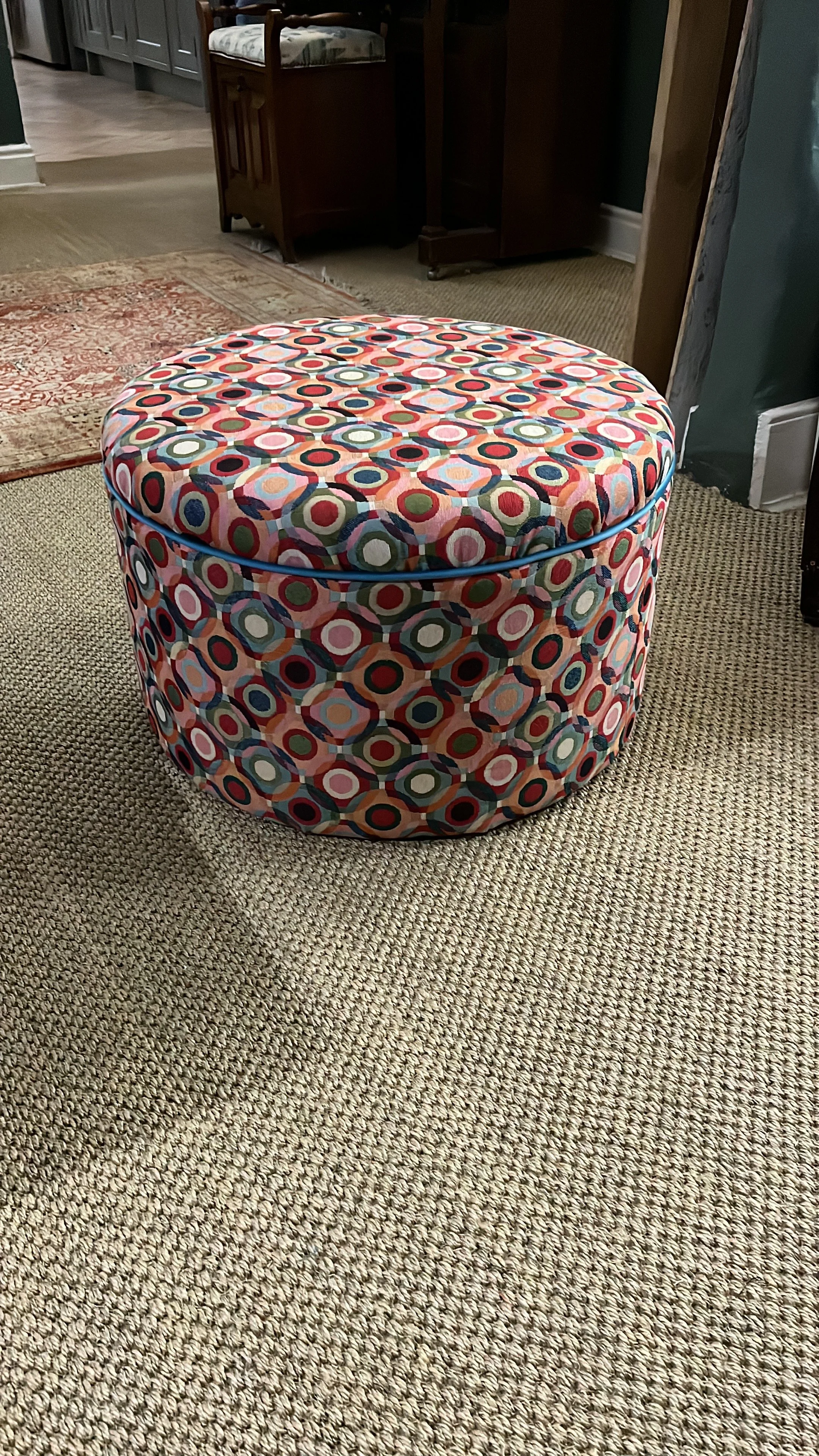 A round upholstered ottoman with a colorful geometric pattern featuring circles and rings in red, blue, white, black, and green, placed on a beige textured carpet in a room with furniture and a doorway in the background.