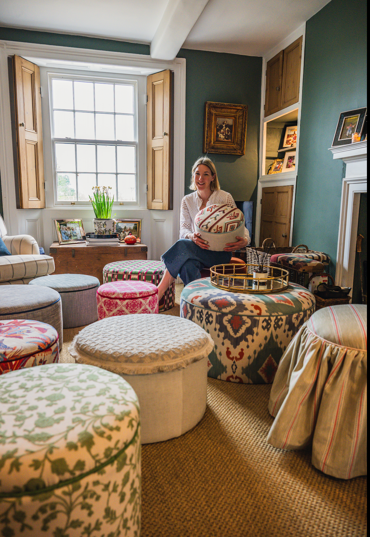 A woman sitting on a colorful upholstered ottoman in a living room, holding a decorative fabric-covered box. The room features patterned ottomans, a fireplace, framed photographs, and a window with wooden shutters.