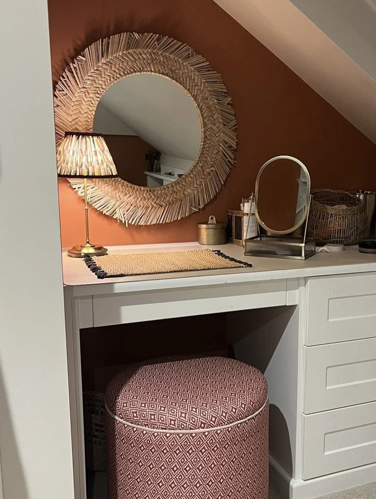 A cozy corner with a round, patterned ottoman, a small white desk with a mirror, a table lamp, and a large decorative round mirror on a warm-colored wall.