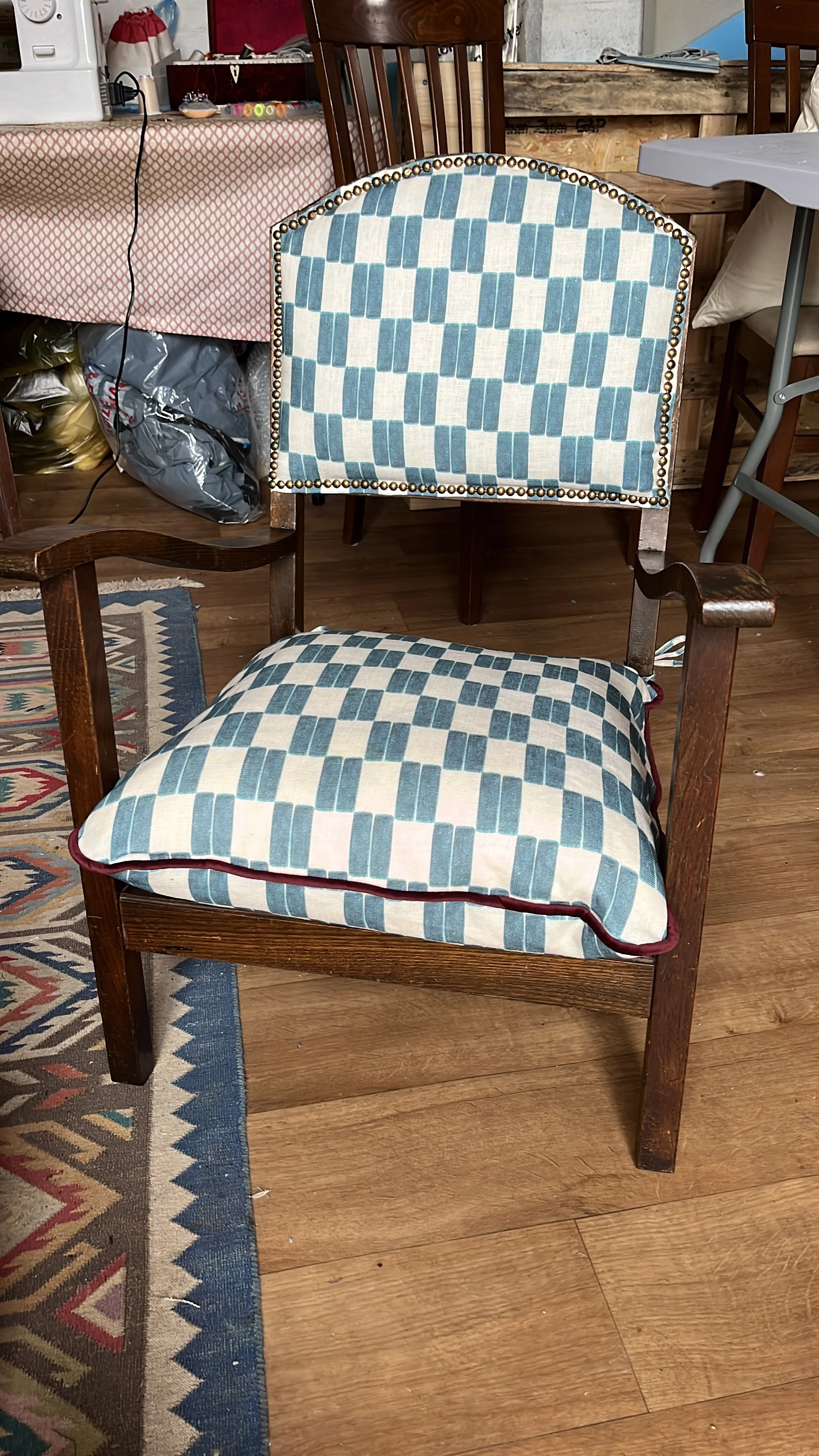 Wooden chair with a cushion on the seat and a matching cushion on the backrest, both with a blue and beige checkered pattern, situated on a wooden floor in a room with various furniture and objects in the background.