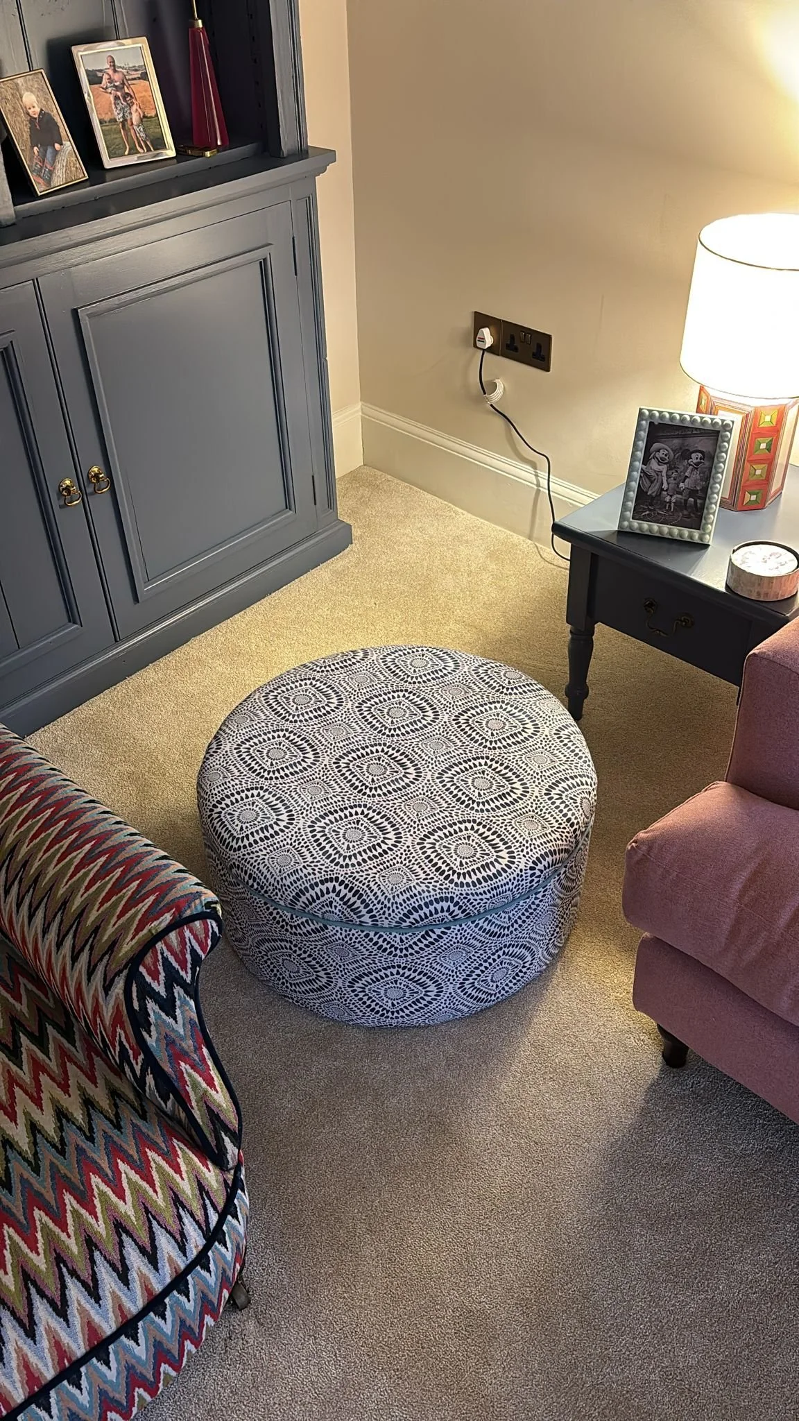 Living room corner with a patterned round ottoman, a pink armchair, and a side table with a black and white framed photo, a lamp, and a decorative item. There is a built-in gray cabinet on the left and an electrical outlet on the wall.