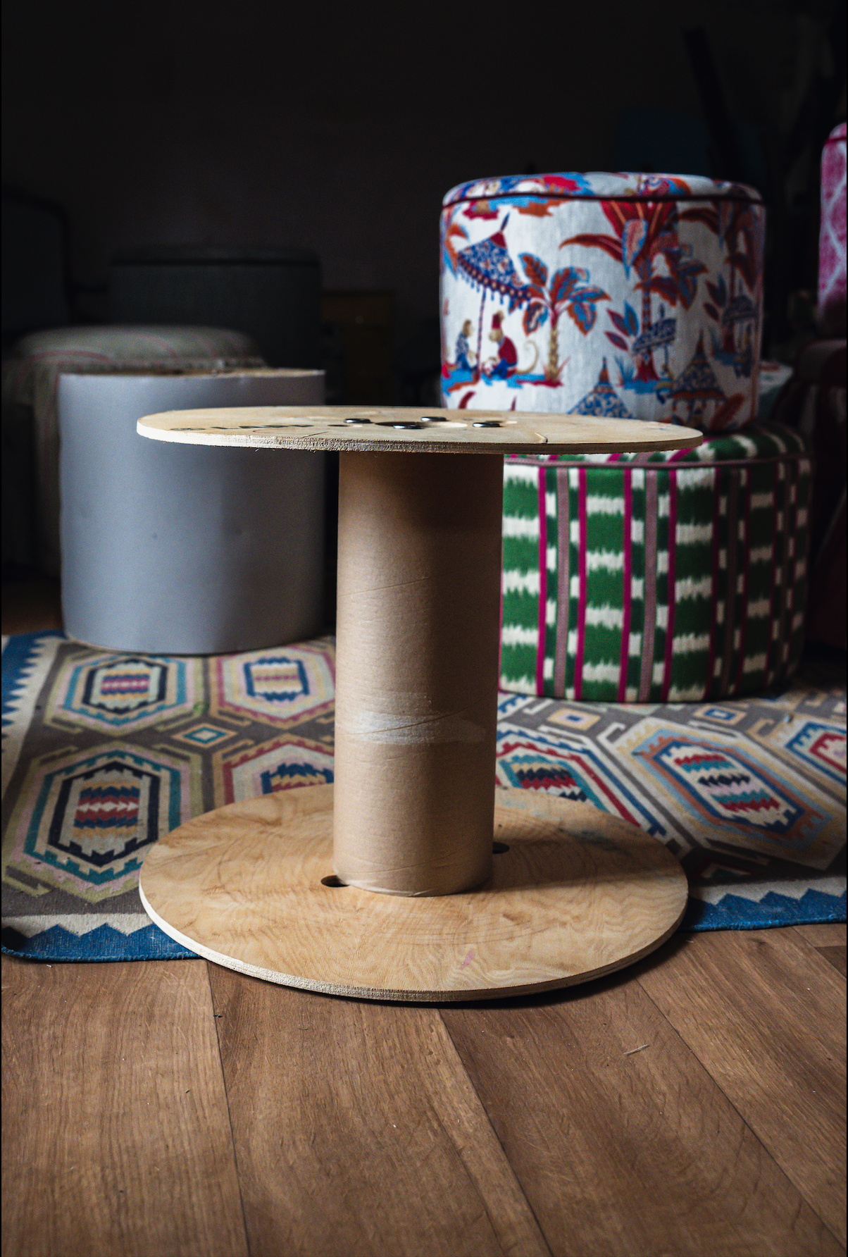 A cardboard dizzy spool table in front of colorful upholstered seats with patterned fabrics. The floor is hardwood, and the background is dark with no distinct objects visible.