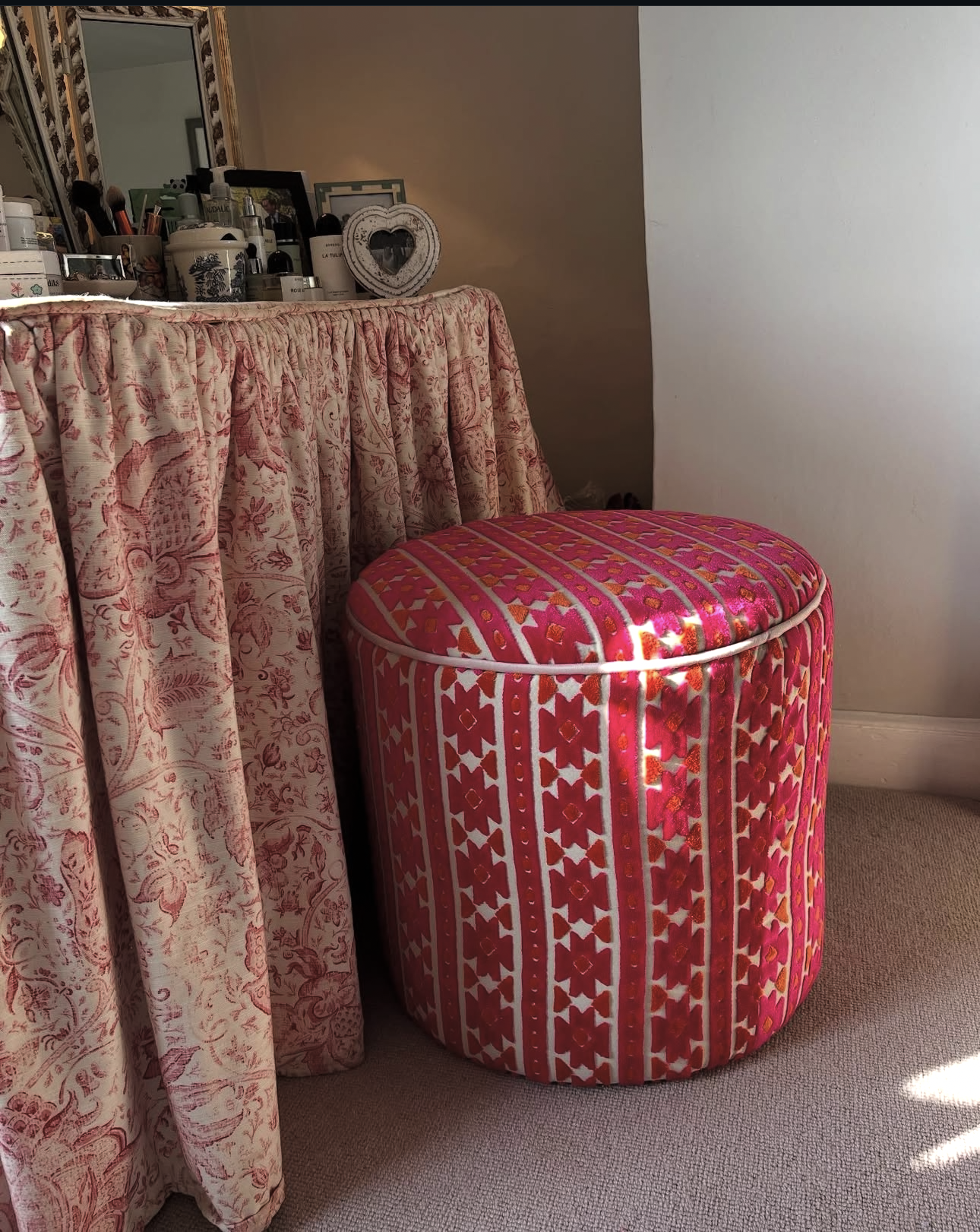 Pink patterned upholstered ottoman next to a floral fabric-covered dresser with various decorative items on top.