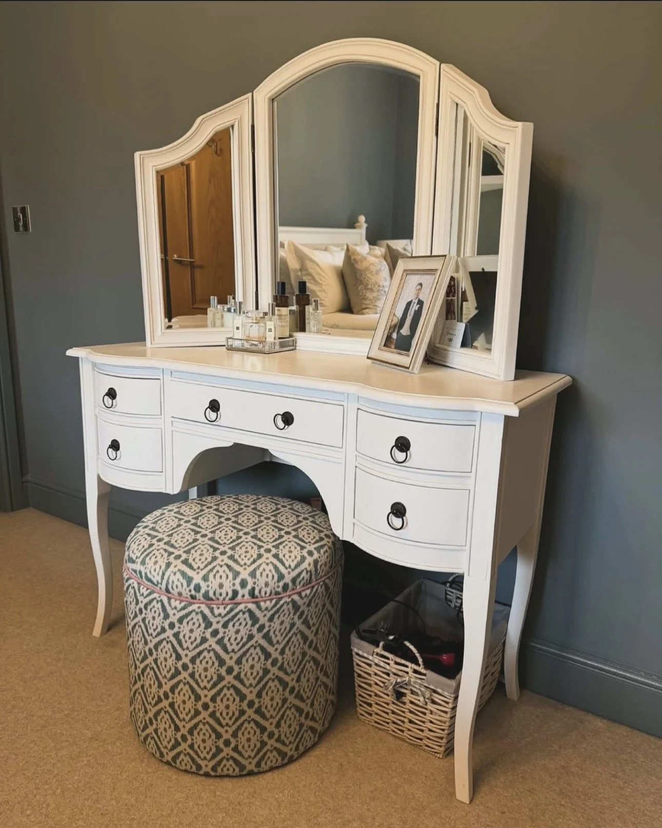 White vanity table with a large mirror, bottles of skincare or perfume, framed photograph, and a small mirror, with an upholstered stool underneath, against a blue wall in a bedroom.