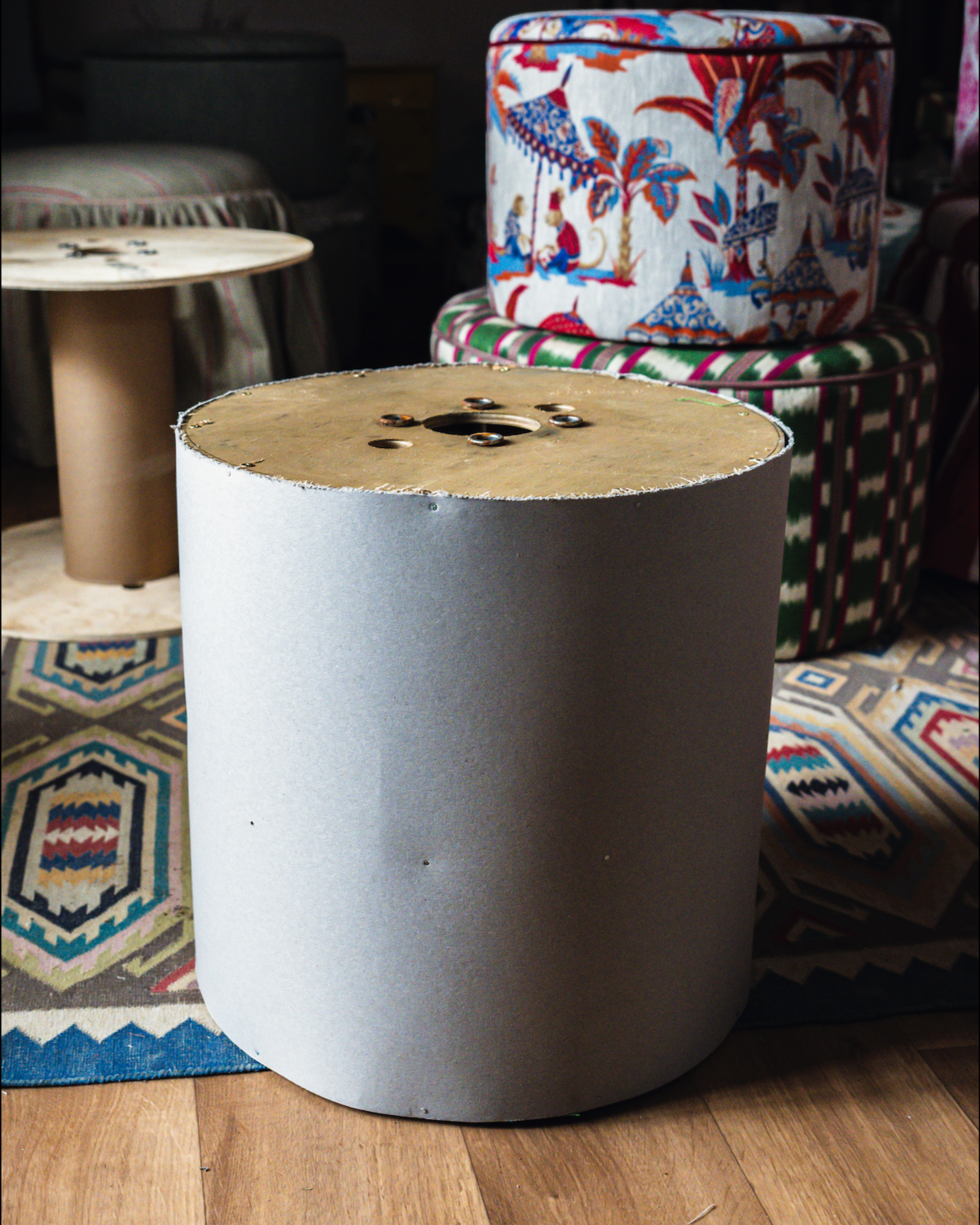 Unfinished cylindrical lampshade with a fabric top featuring a decorative design of children and animals, placed on a patterned rug in a room with furniture and other lampshades.