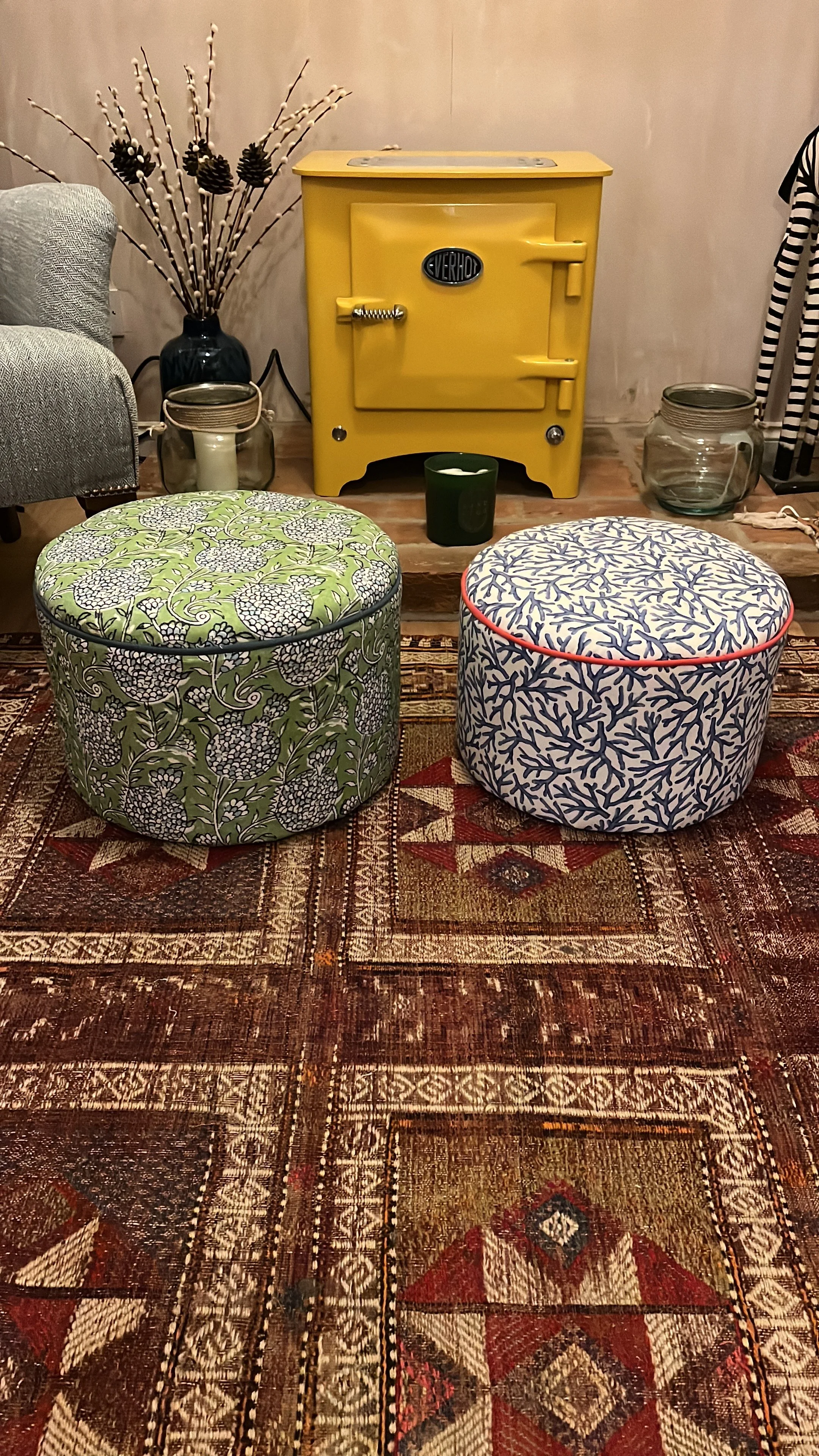 A living room scene featuring a yellow oven, two round patterned ottomans on a large oriental rug, and decorativev items including a vintage glass jar, a candle, and a vase with dried branches and pine cones.