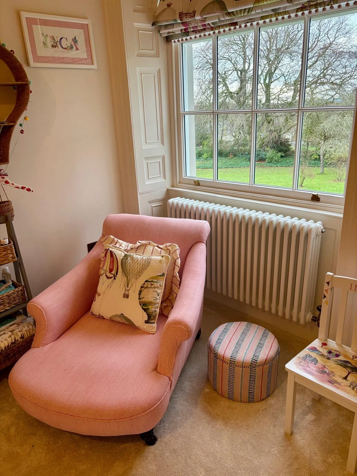 A cozy sitting area by a large window showing an outdoor garden with bare trees and green grass. The space includes a pink upholstered chaise lounge with a decorative pillow, a striped round ottoman, a small white table with a colorful butterfly and 