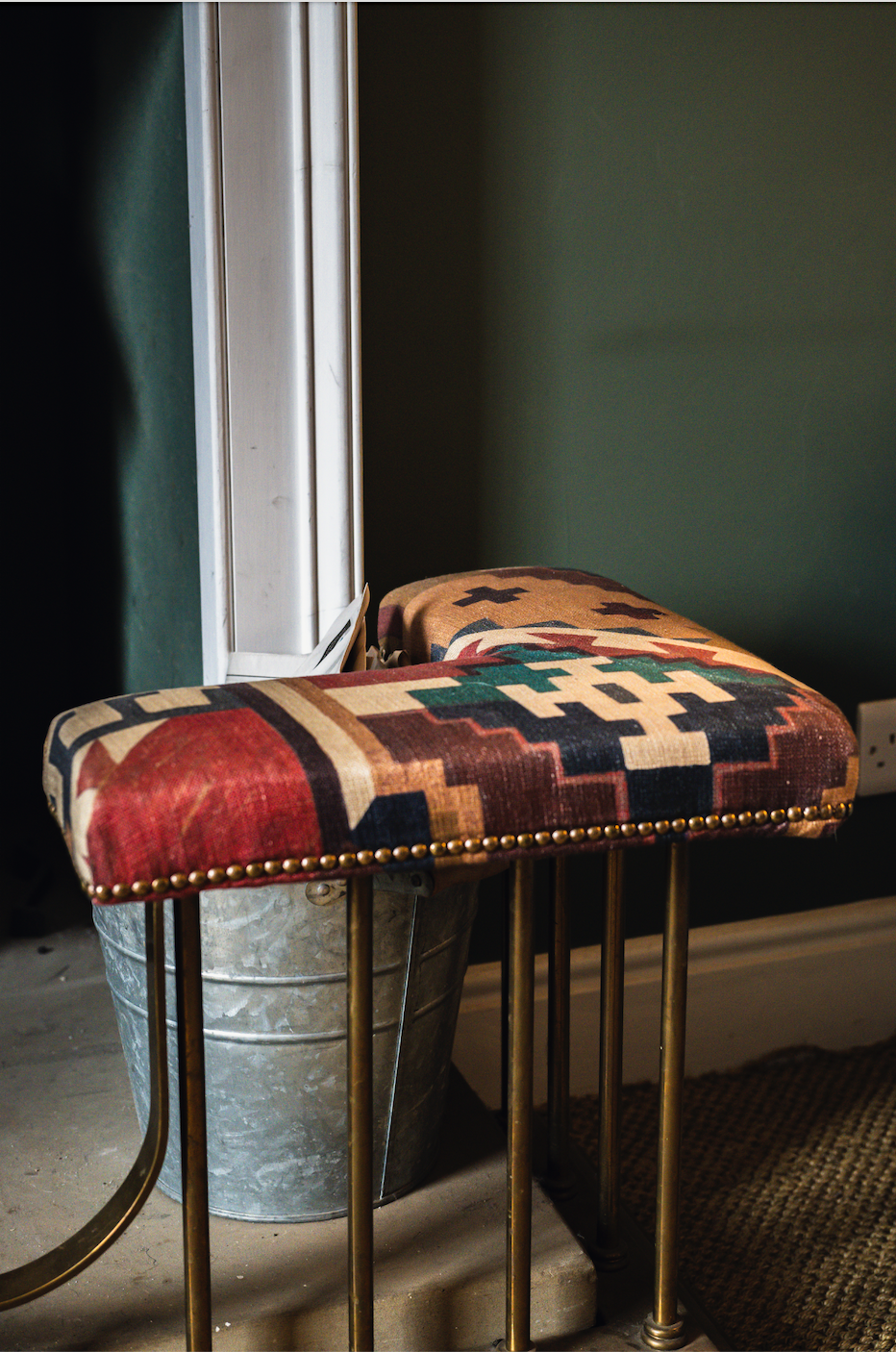 A decorative stool with a colorful geometric patterned fabric top, supported by gold-colored metal legs, placed next to a metal bucket. The setting appears to be indoors near a wall with a baseboard.