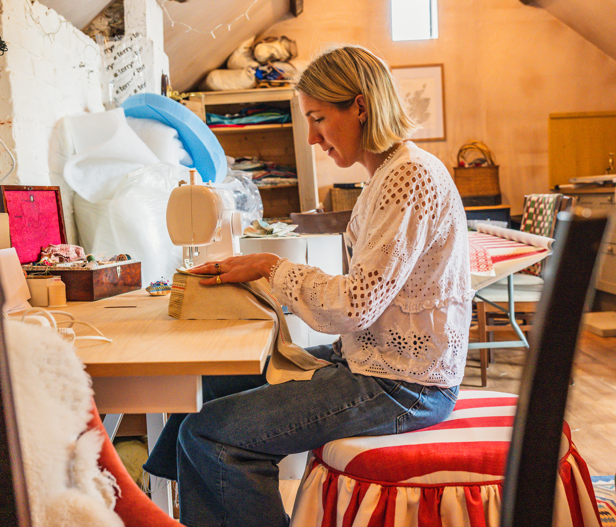 A woman with blonde hair sewing at a desk in a cozy, cluttered room. She is wearing a white eyelet blouse, and there are various sewing supplies and fabrics around her. The room has warm lighting, with a sloped ceiling and wooden shelves.
