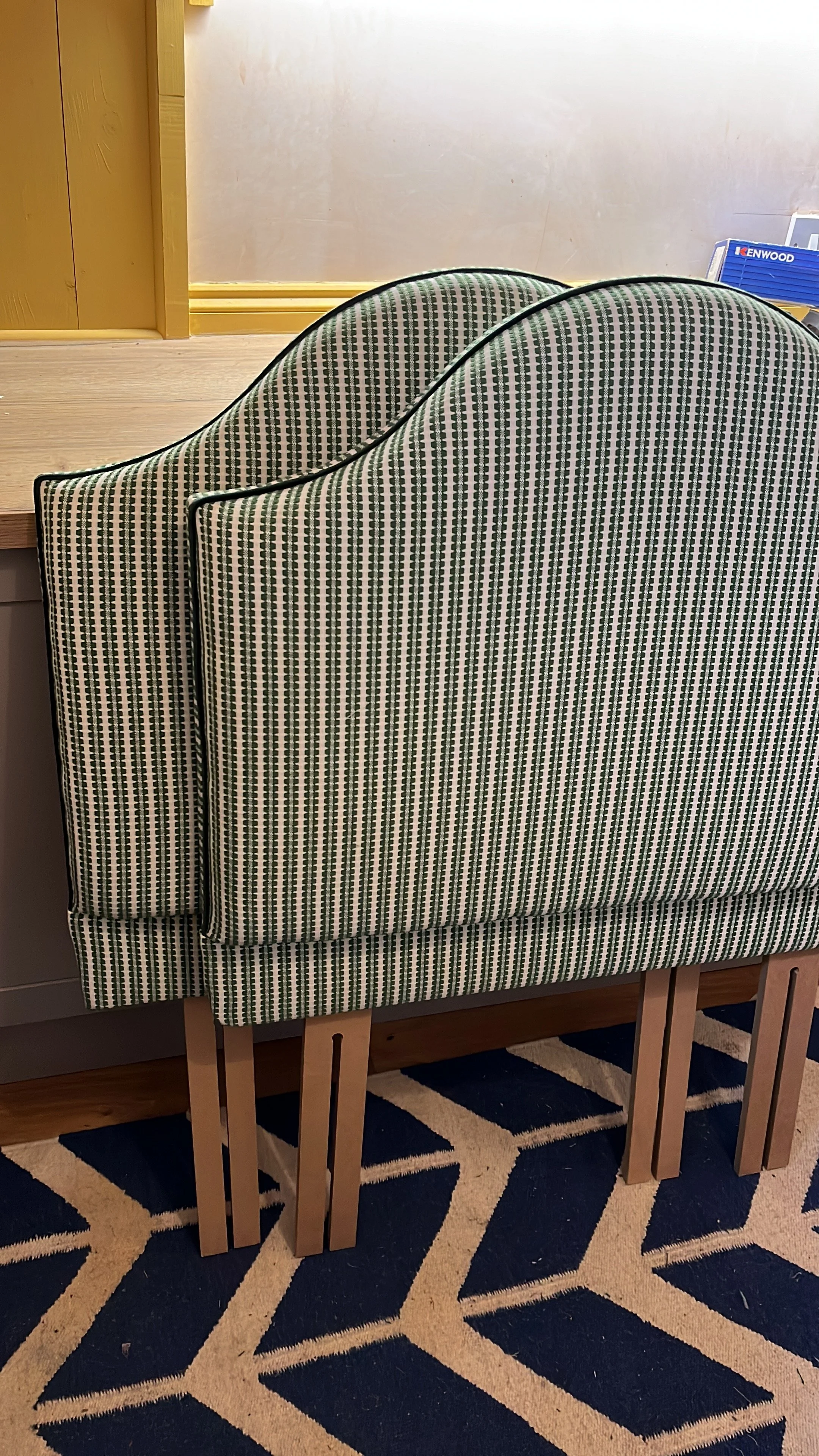 Two patterned chairs stacked together with wooden table legs visible underneath and a blue tile patterned rug beneath, in a room with a light-colored wall and a wooden counter or table behind them