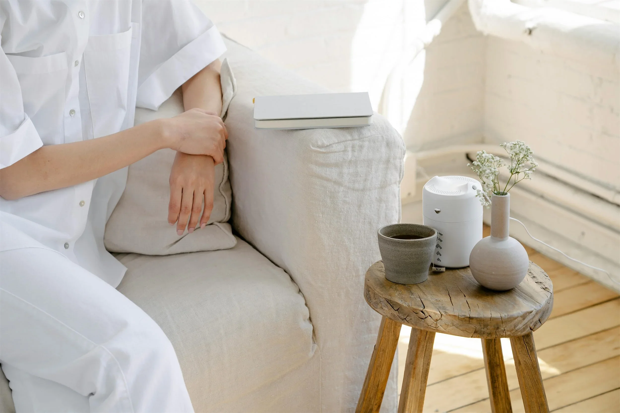 A cozy corner with a white sofa, a small round wooden table, a white diffuser, a ceramic cup with a hot beverage, and a vase of white flowers. A person in a white shirt is resting their arm on the sofa.
