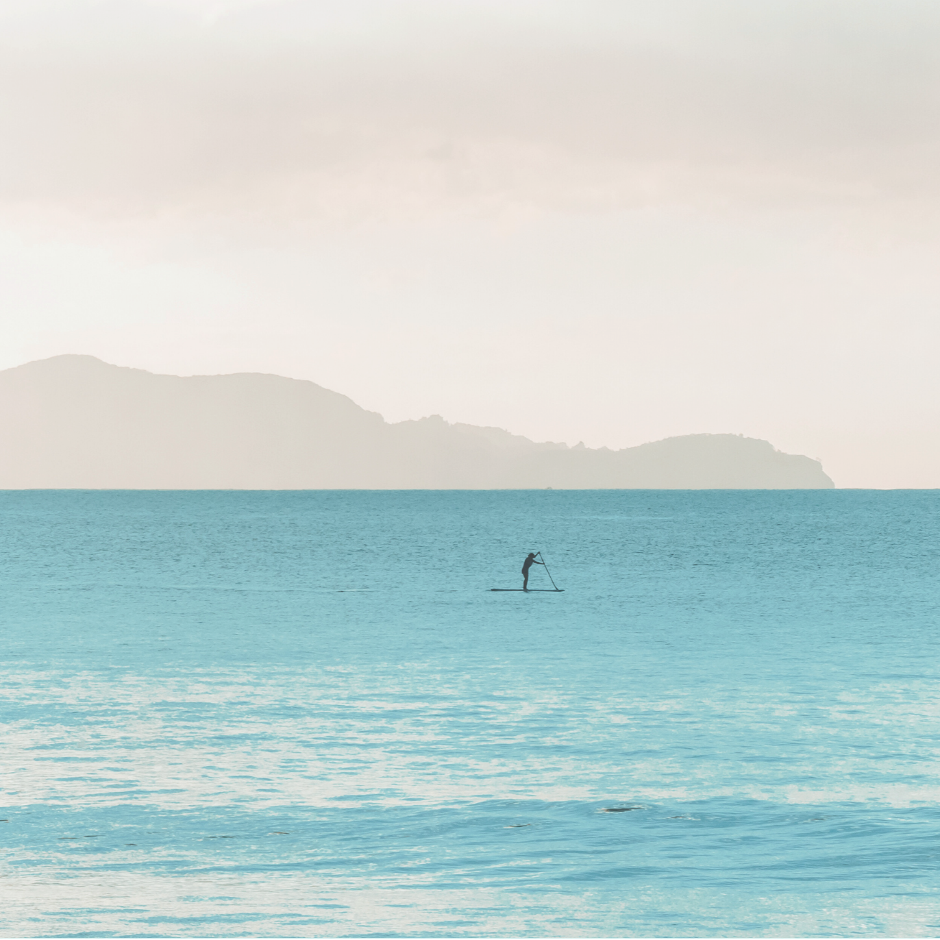 A lone paddleboarder on calm open water with misty mountains in the background, symbolising individual journey and inner calm.