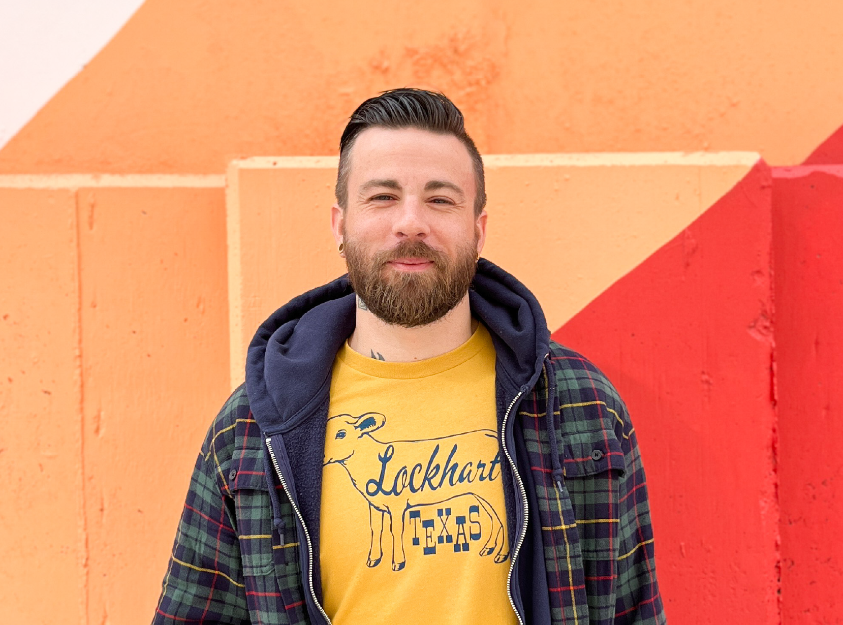 A man with dark hair and a beard standing in front of an orange wall with red and cream geometric patterns. He is wearing a yellow T-shirt with a cow graphic and the word "Lockhart" and "Texas," layered with a blue hoodie and a green plaid jacket.