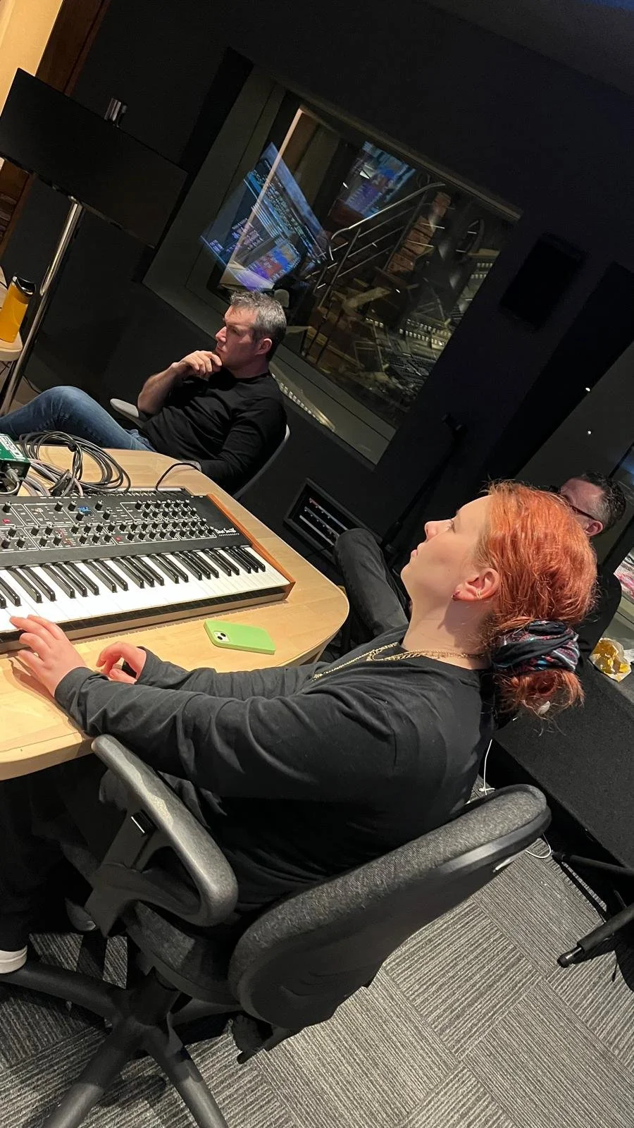 Miranda Joy sitting at a desk with a keyboard synthesizer in a recording studio, with two men in the background, one wearing glasses and the other with short hair, all in a dark room with soundproof walls.