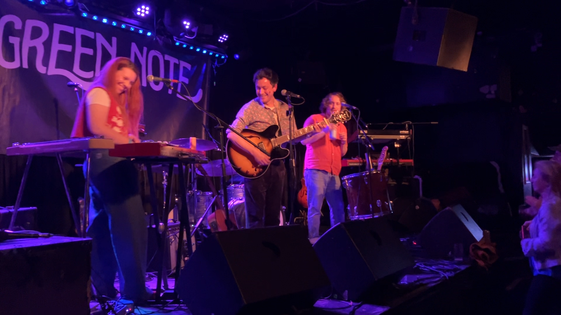 Miranda Joy performing live on stage at a concert, with three musicians playing instruments and singing, Green Note sign in background.