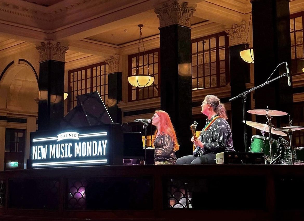 Two women performing music on stage at a venue with a sign that reads "NEW MUSIC MONDAY". One woman is playing the piano, and the other is playing the guitar.