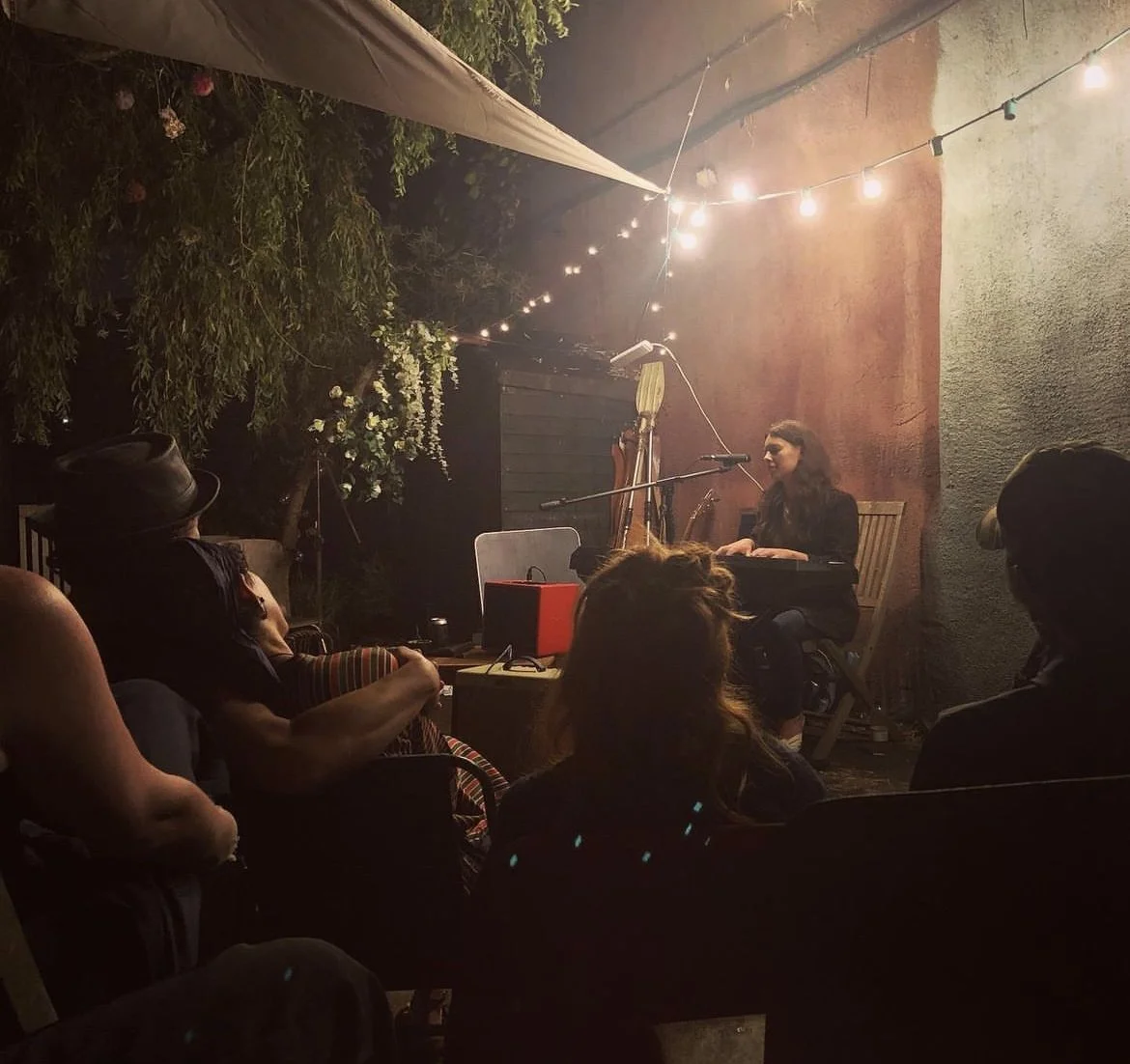 Miranda Joy playing keyboard and singing into a microphone during an outdoor nighttime acoustic performance, with string lights overhead and audience members seated and listening.