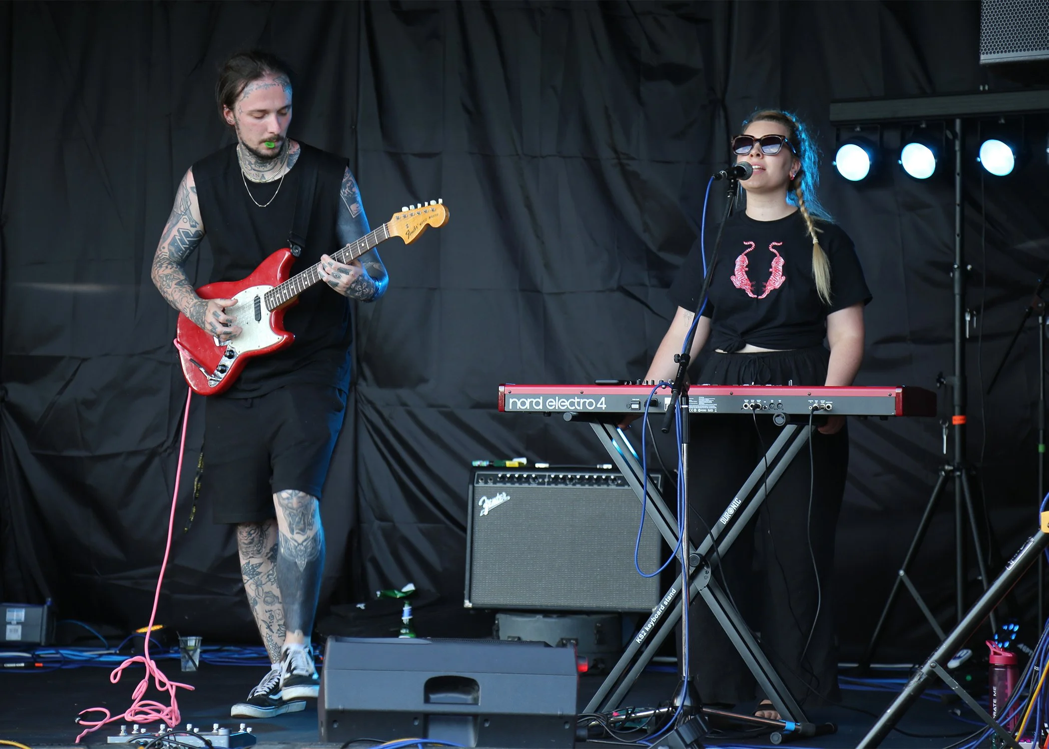 A male guitaristplaying an electric guitar and Miranda Joy singing into a microphone on stage during a live performance, with stage lights and black backdrop.